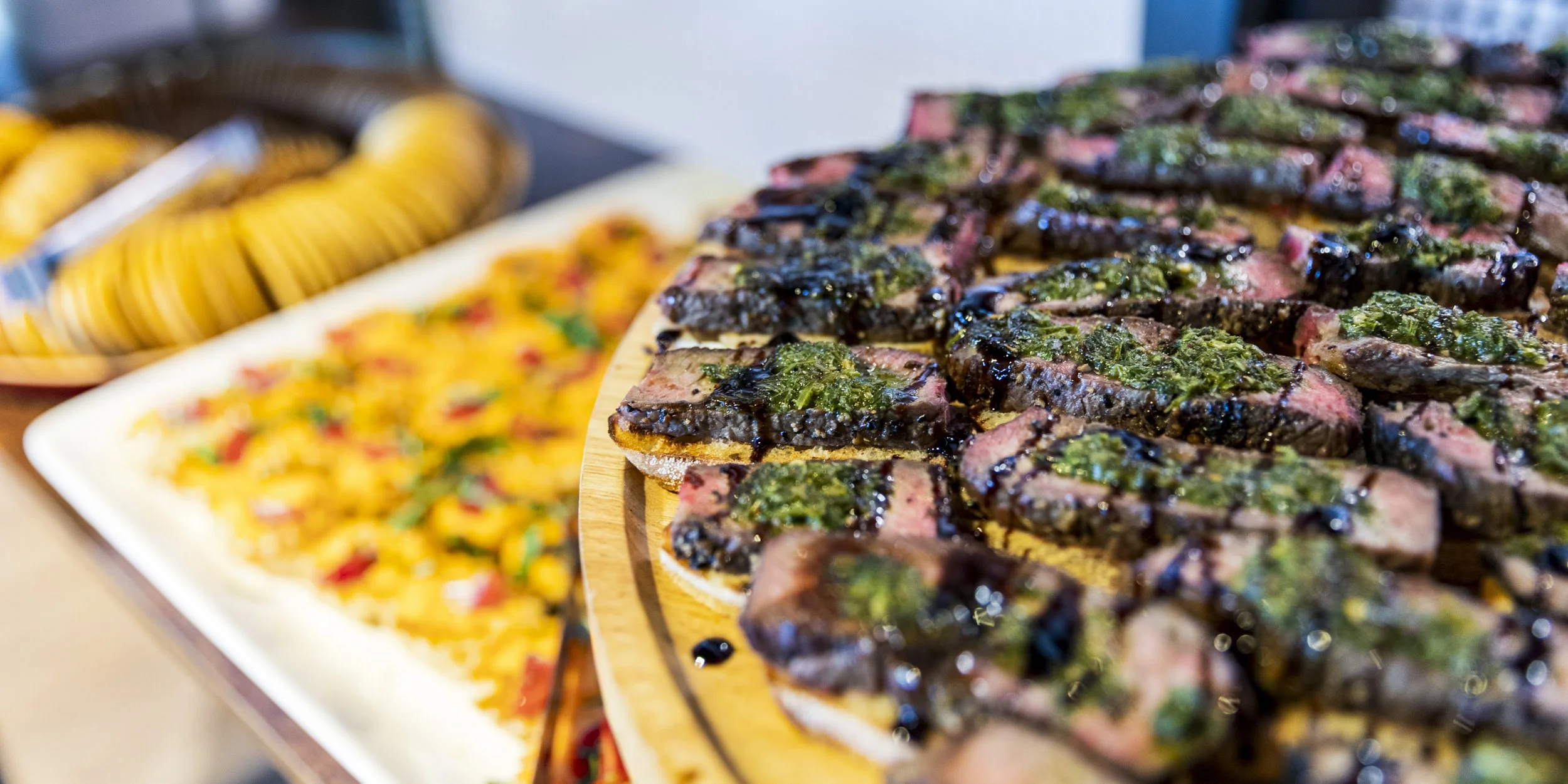 Close-up of sliced steak topped with green herbs and sauce on a round wooden platter, with other food dishes in the background.