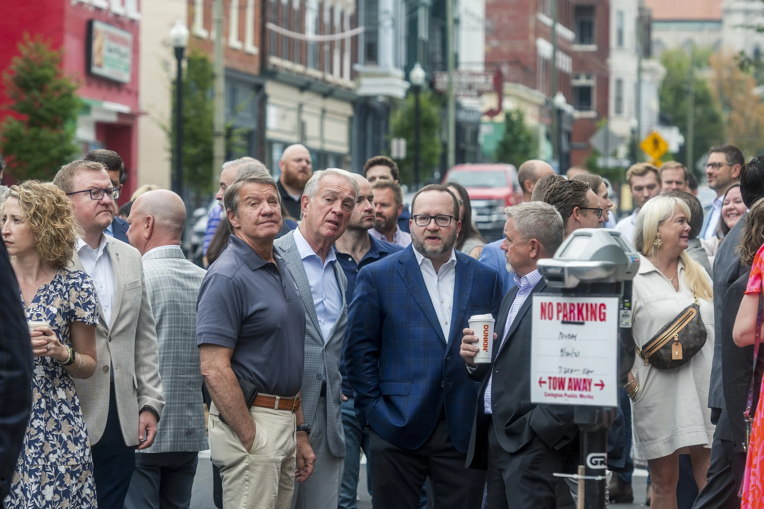 A crowd of people gathered on a city street, engaged in conversation, with buildings and cars in the background, and a no parking sign in the foreground.