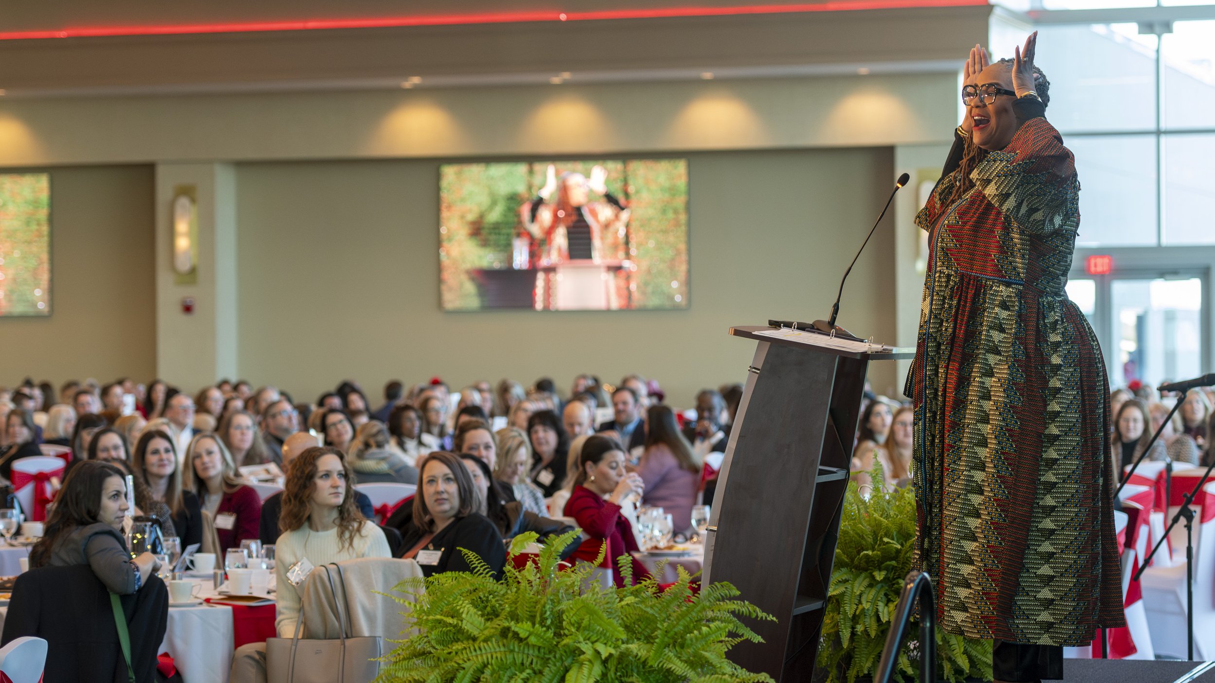 A woman in a colorful dress speaking at a podium during a large conference, with an audience seated at round tables in a spacious, well-lit room.