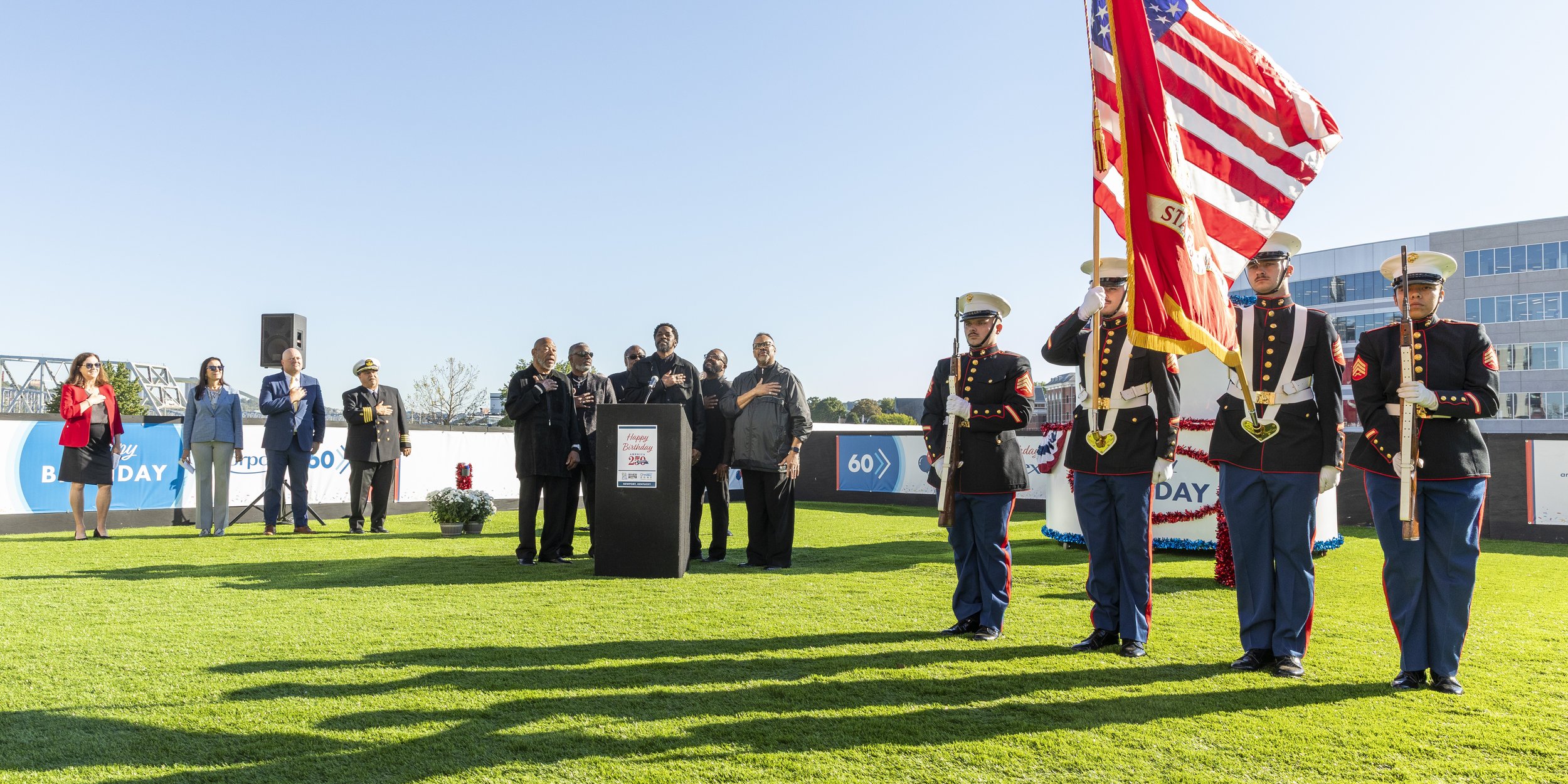A group of people, including firefighters and officials, standing on a grass field during a ceremony with a flag being presented, with a building and clear sky in the background.