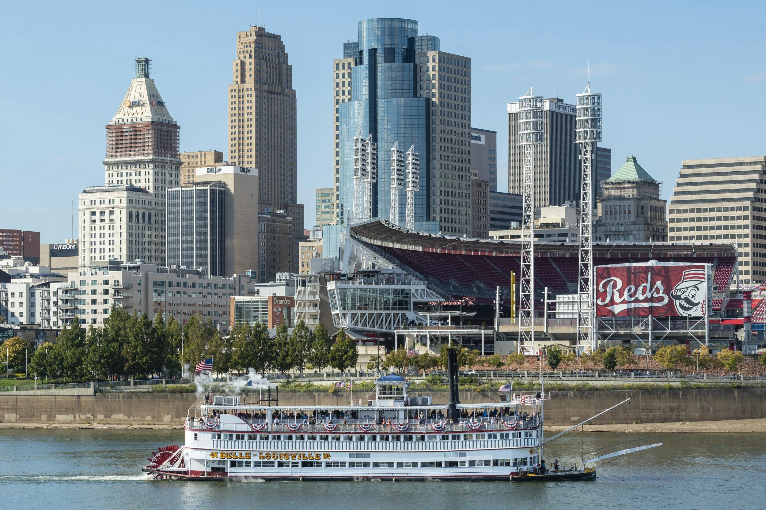 A riverboat named Belle of Louisville sailing on a river with a city skyline in the background, including high-rise buildings and a stadium with a large Cincinnati Reds banner.