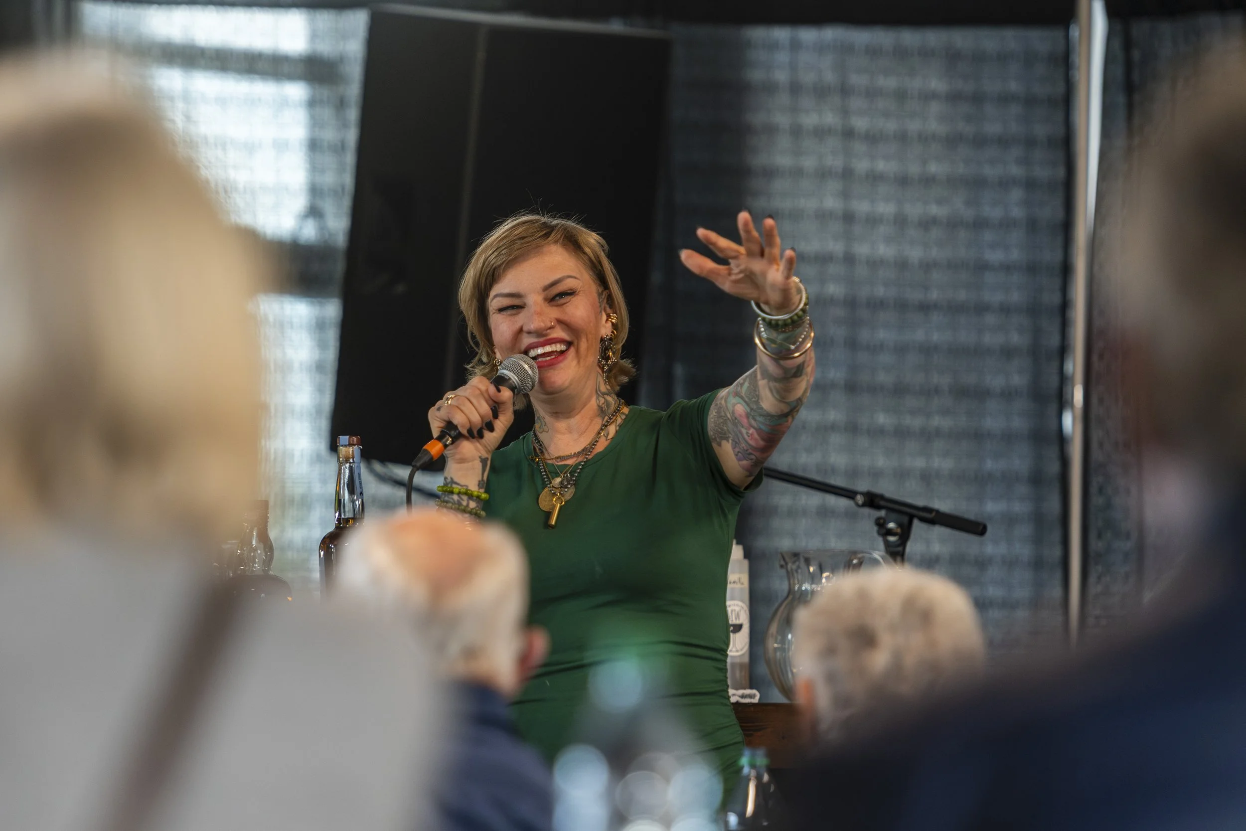 A woman with short blonde hair and tattoos on her right arm, smiling and speaking into a microphone, at a public event or performance.