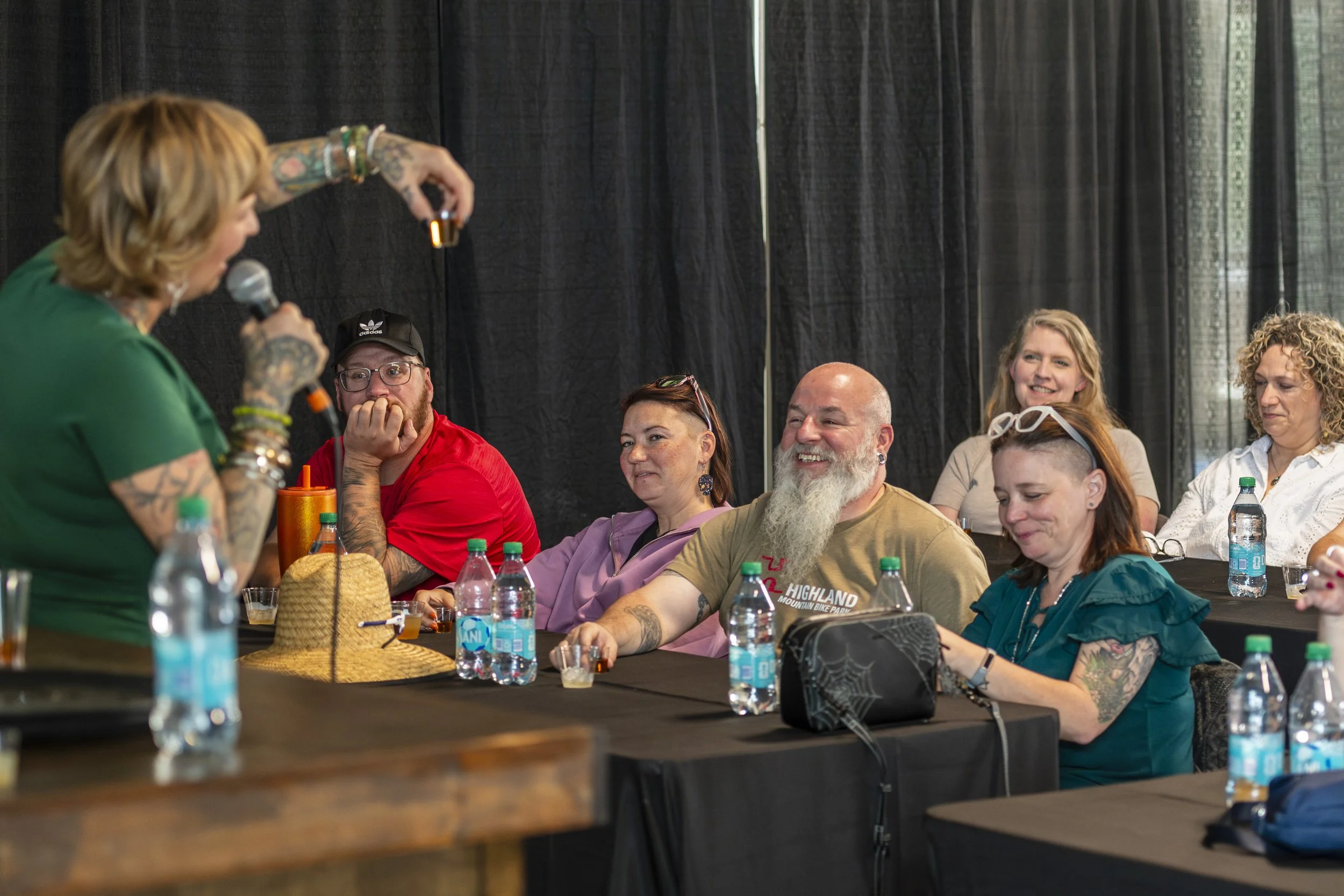 Woman with tattoos speaking into a microphone at a table with several people listening and smiling, with water bottles on the table.
