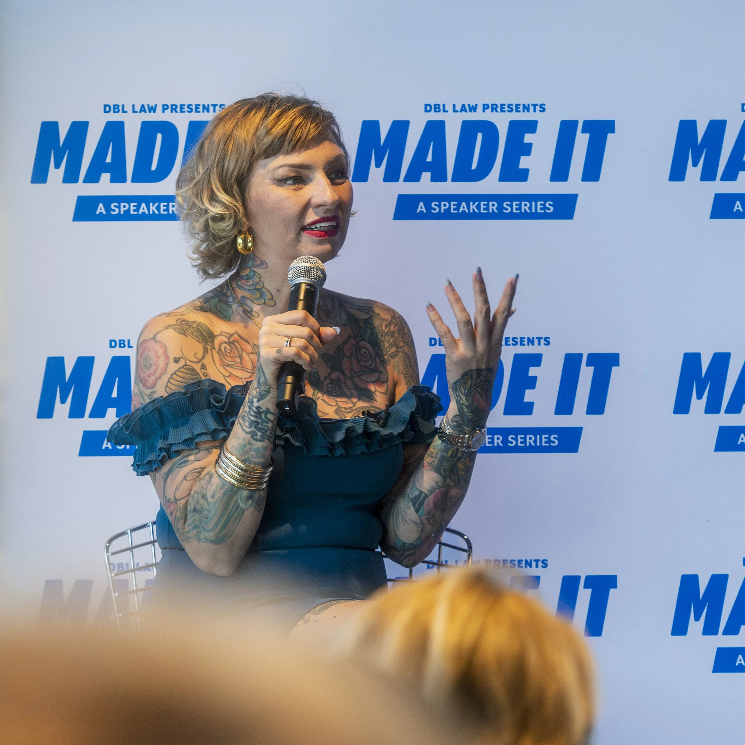 Woman with tattoos, short blonde hair, wearing a dark blue off-the-shoulder dress with ruffled sleeves, speaking into a microphone at a speaker event, with a backdrop reading "Made It" and "A Speaker Series".