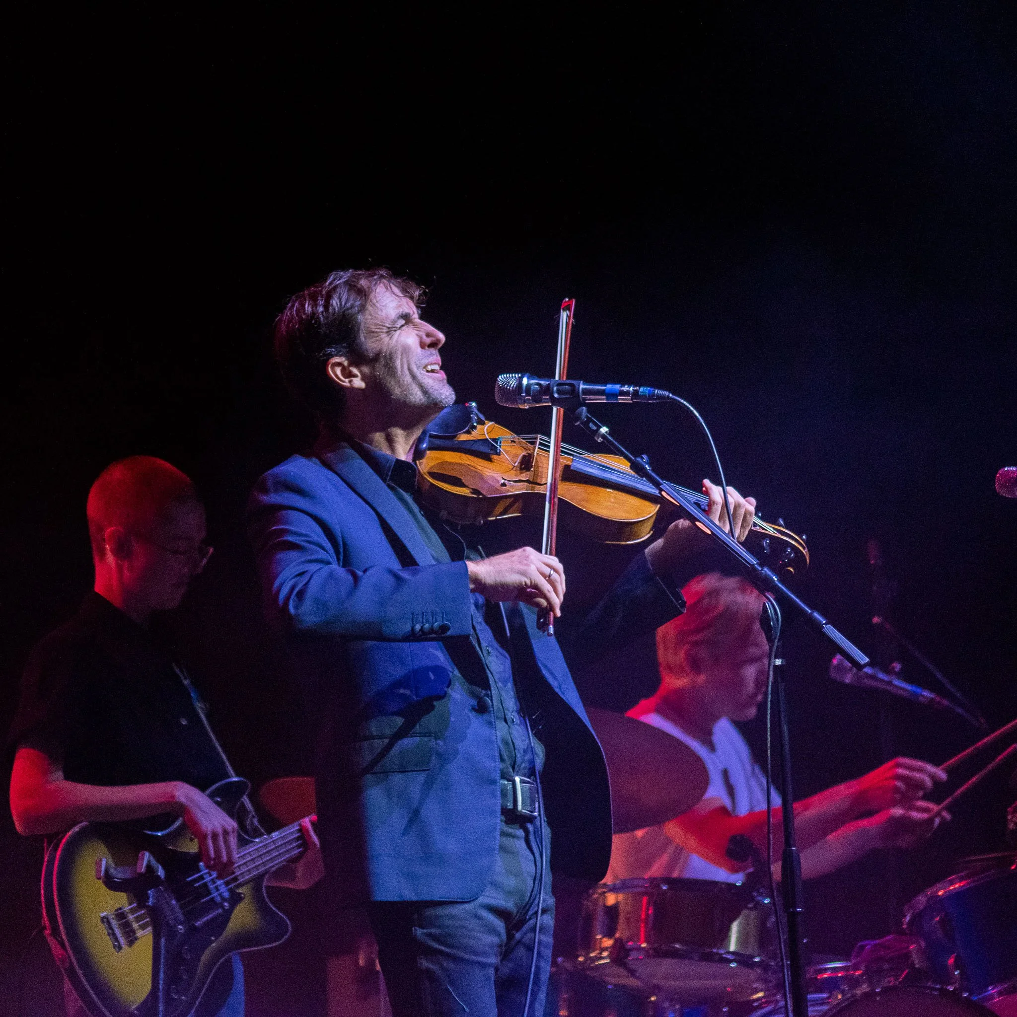 A man playing violin sings into a microphone during a live music performance, accompanied by band members playing guitar and drums in a dimly lit concert setting.