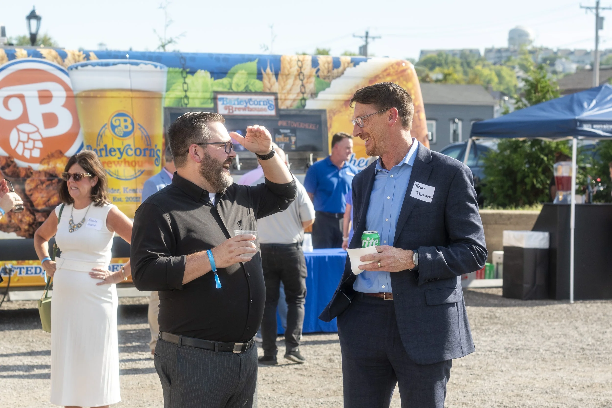 Two men are smiling and talking at an outdoor event, holding drinks, with a blue canopy tent and a large colorful sign in the background.