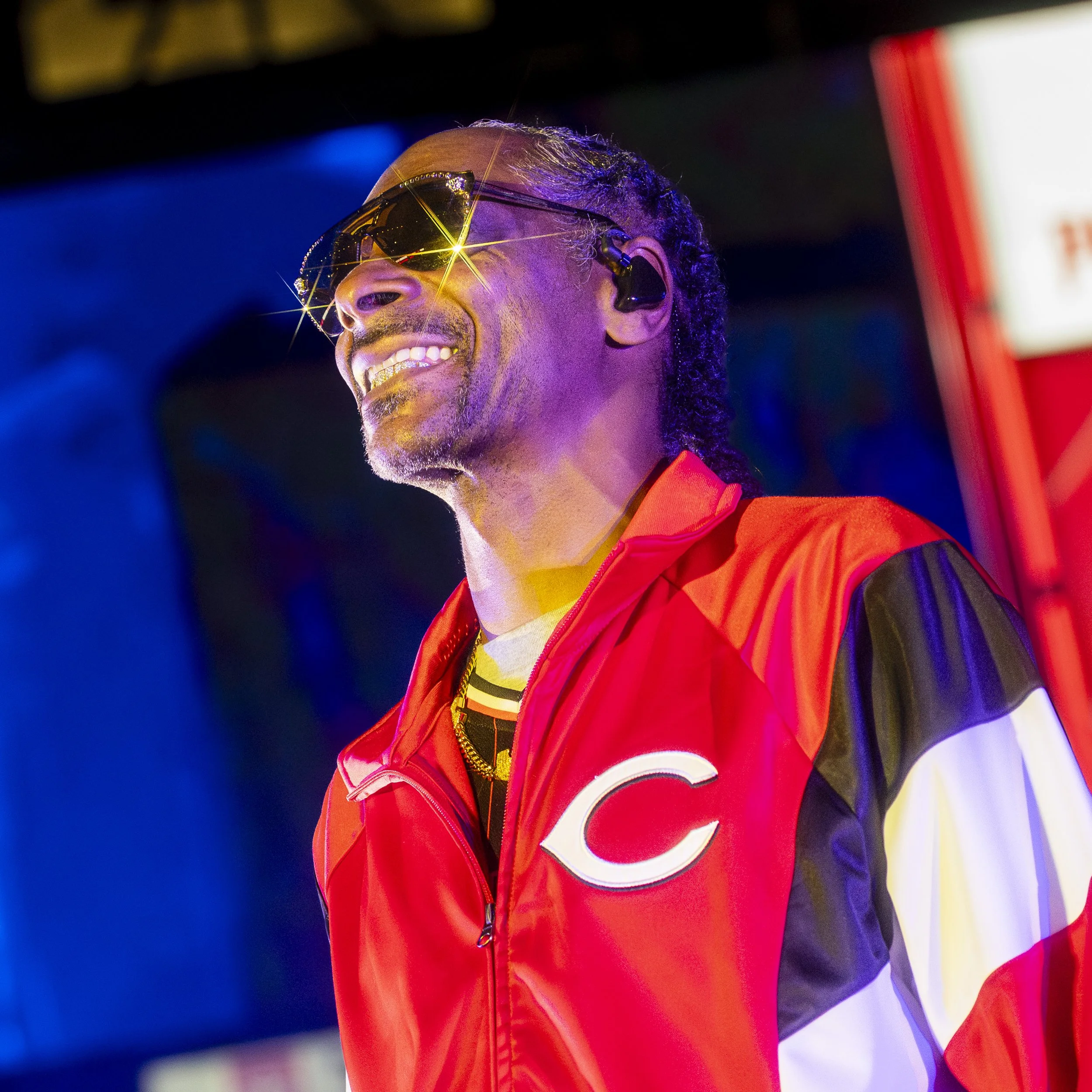 A man smiling, wearing sunglasses with a starburst reflection, a red and black jacket with a white 'C' logo, and in-ear monitors, in a warmly lit environment.