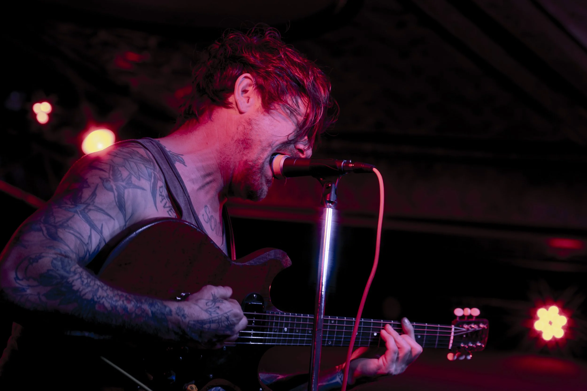 A man with tattoos on his arms and chest, singing into a microphone while playing an acoustic guitar, with red stage lighting in the background.