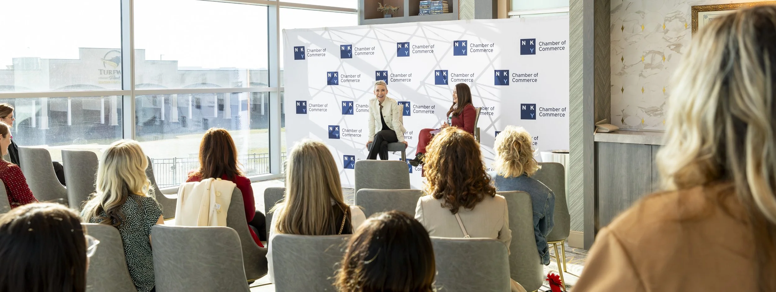 Two women speaking on stage during a panel discussion at a Chamber of Commerce event, with seated audience members listening. The stage backdrop displays the Chamber of Commerce logo.