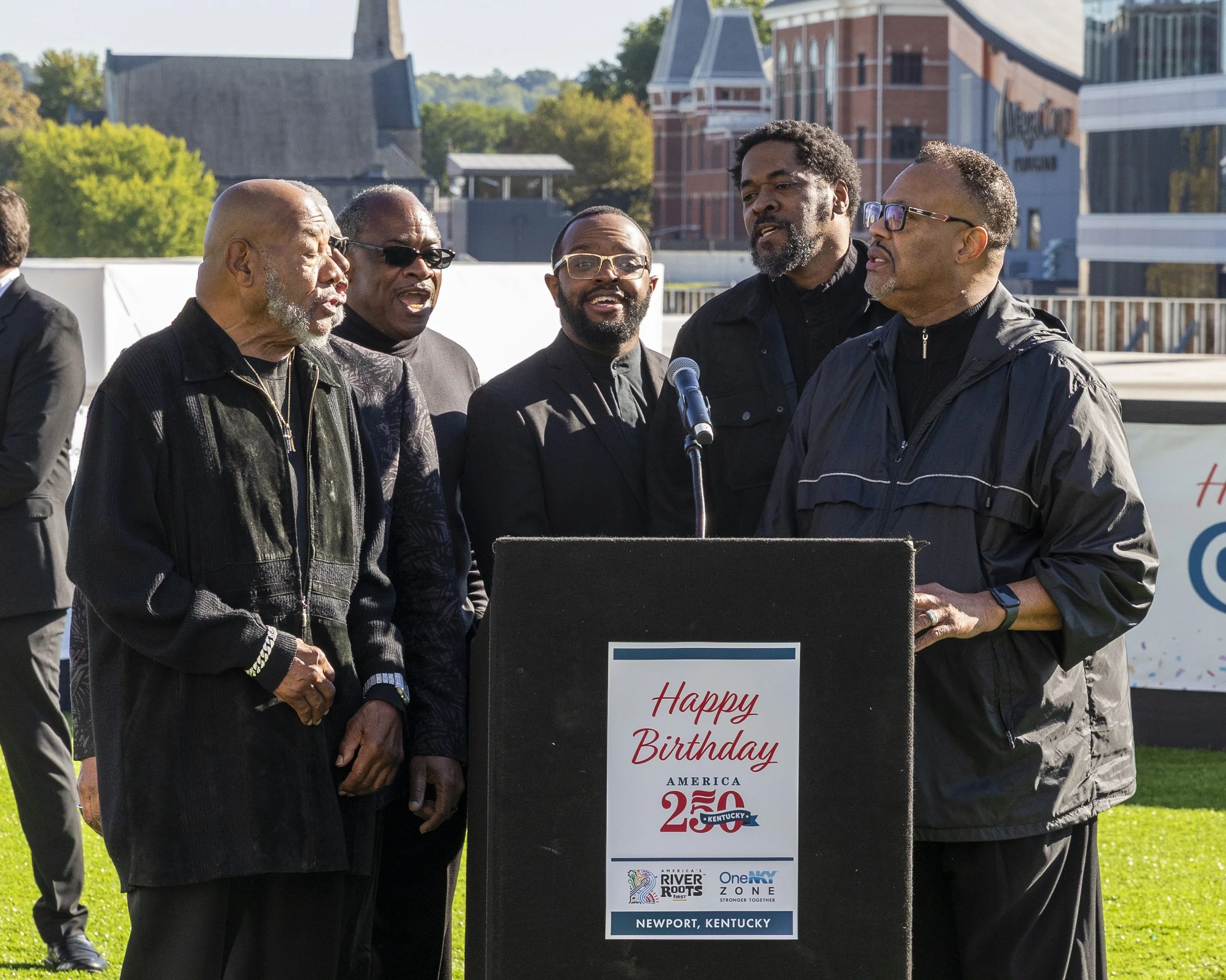 A group of six men standing outdoors, with a podium that says 'Happy Birthday America 250 Kentucky' and the words 'Newport, Kentucky' at the bottom. They are dressed in mostly dark clothing, with some wearing glasses, and are gathered around a microp