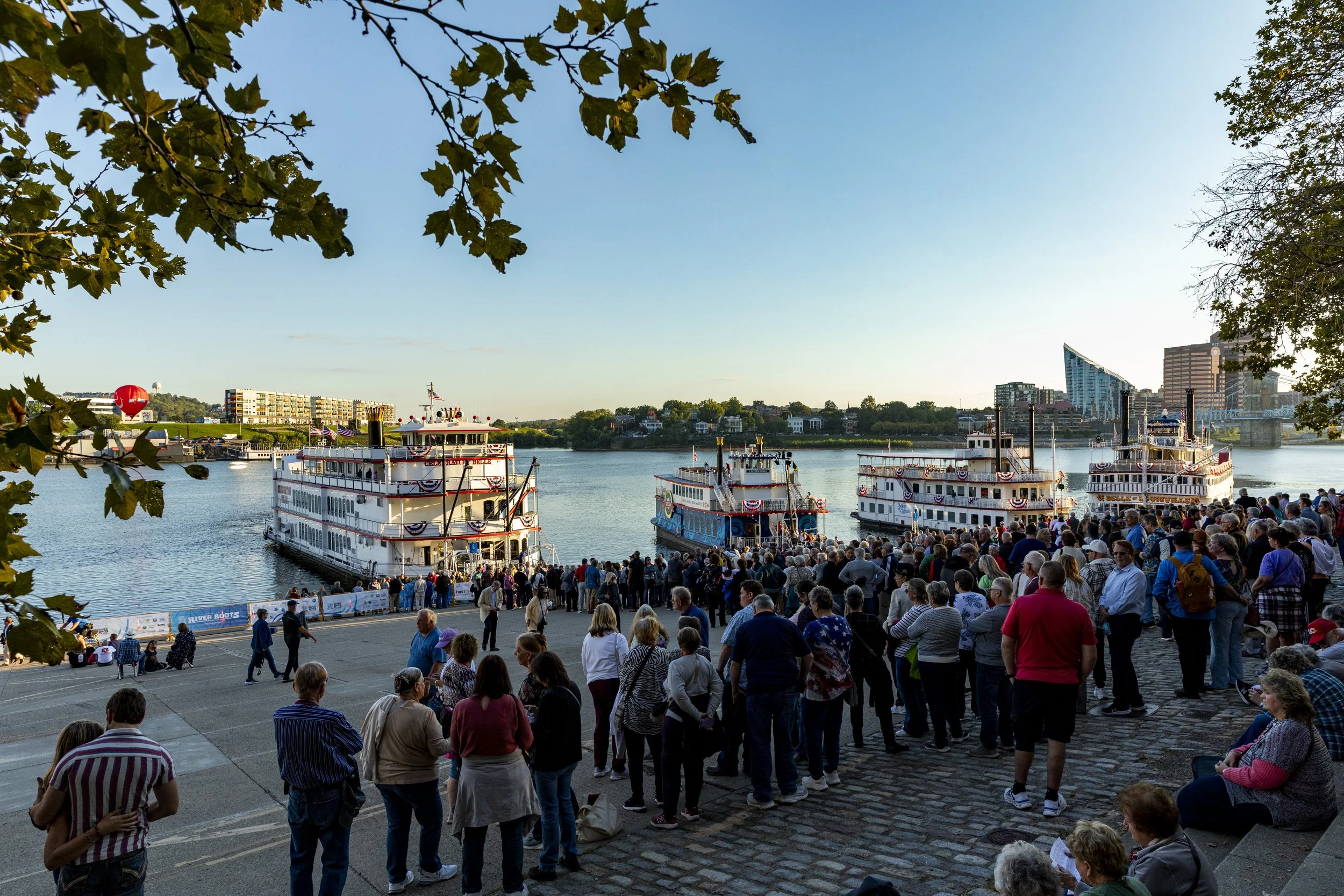 A large crowd of people gathered along the riverbank, waiting to board three paddlewheel boats decorated with patriotic bunting, on a clear day with blue skies and city buildings in the background.