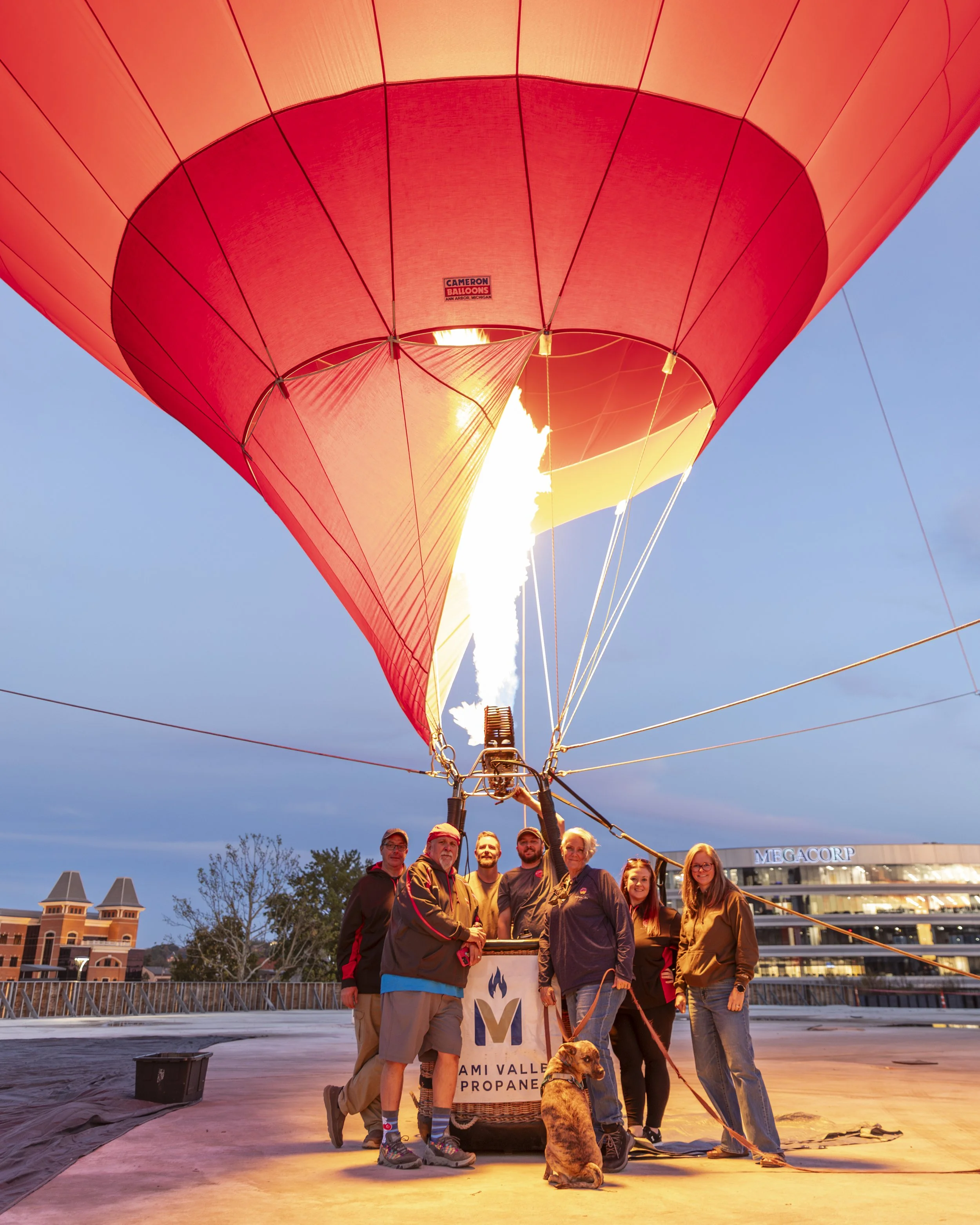 Group of people preparing a hot air balloon for takeoff on a rooftop during dusk, with a dog present, and buildings visible in the background.