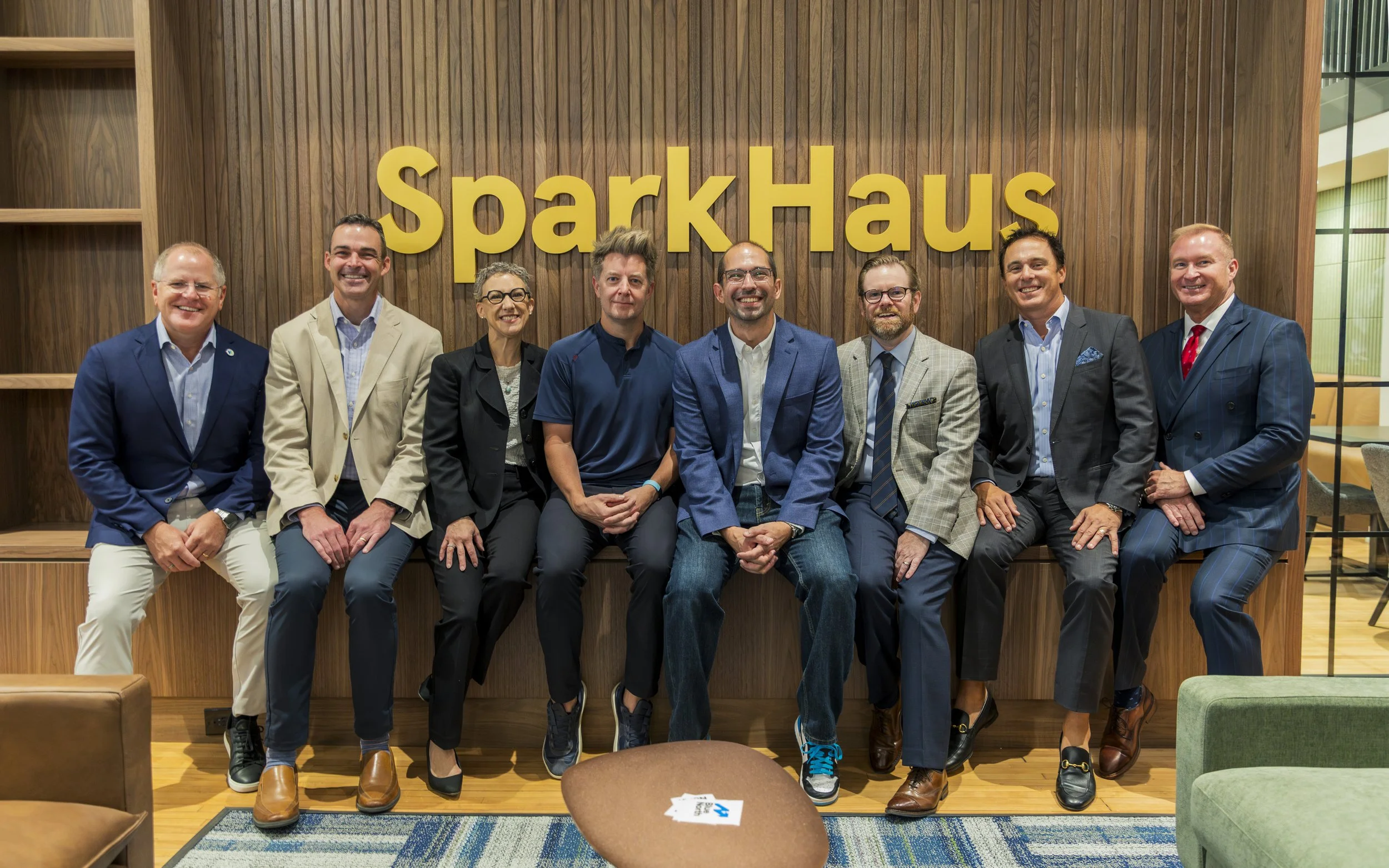 A group of nine professionally dressed individuals sitting on a wooden bench in front of a wood-paneled wall with the large yellow sign 'SparkHaus' in the background.