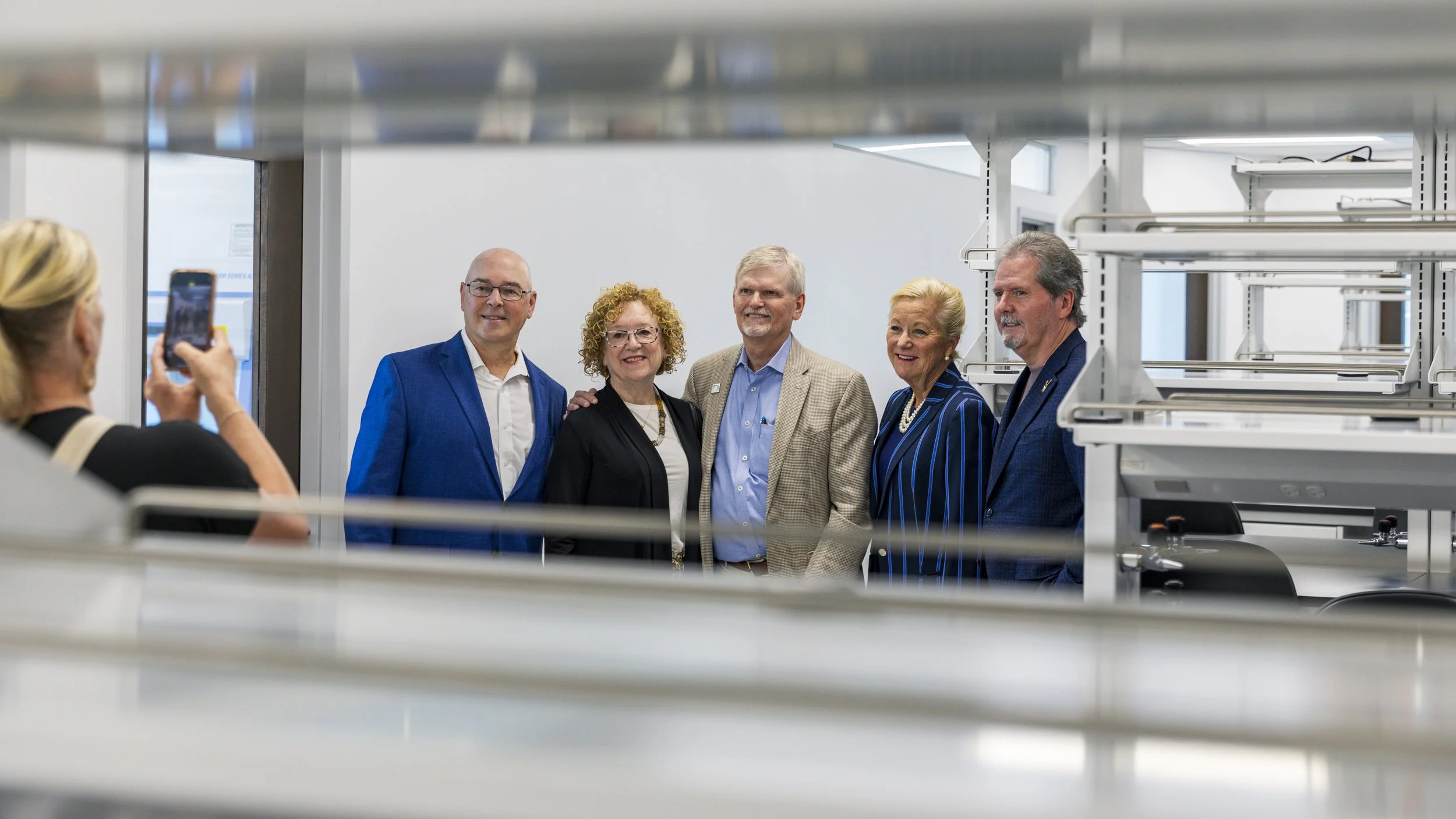 Group of five professionally dressed adults posing for a photo in a modern office or lab environment, with a woman in the foreground taking their picture.