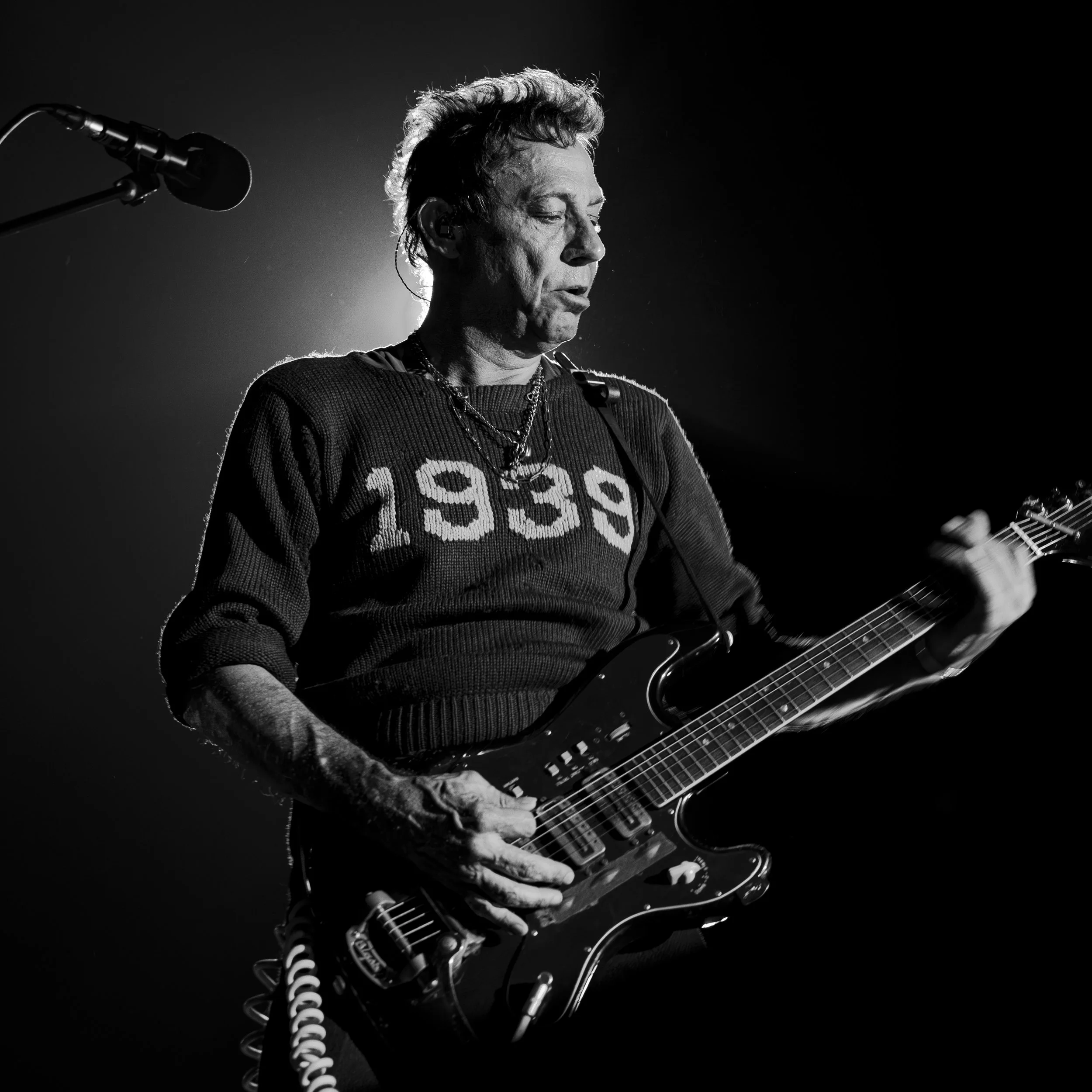 Black and white photo of a male guitarist performing on stage, wearing a sweater with '1939' on it, with a guitar and microphone, under stage lighting.