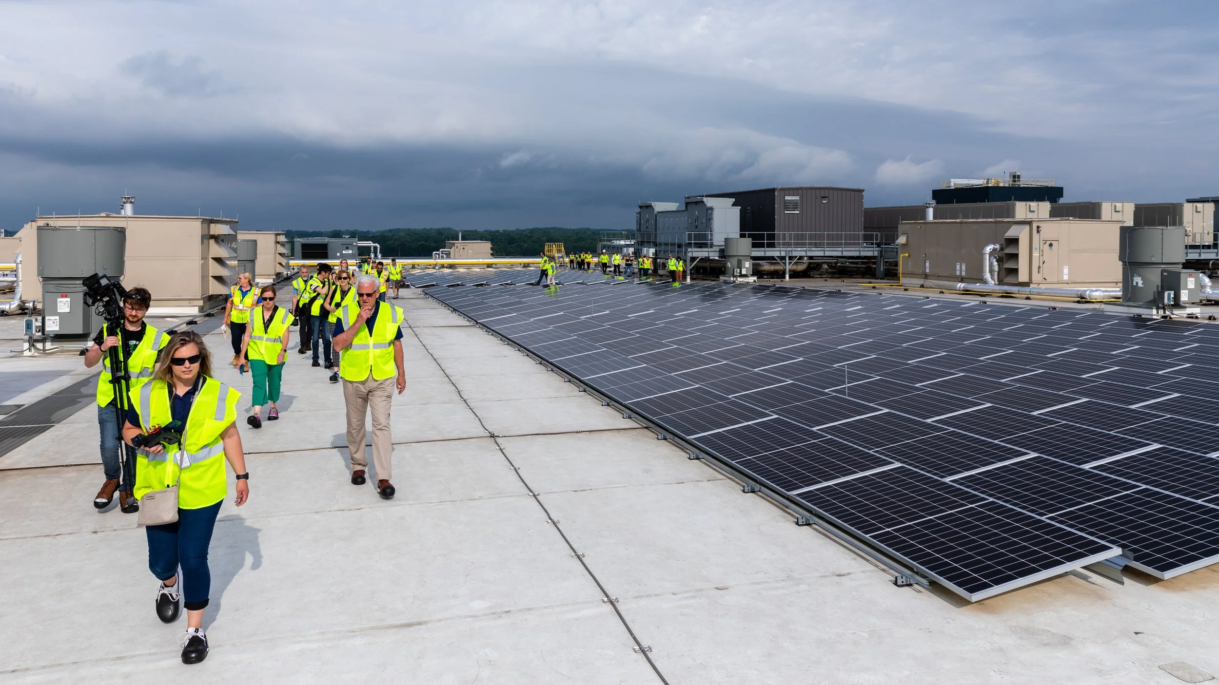 Group of people walking on a rooftop with solar panels, wearing yellow safety vests, under cloudy skies.