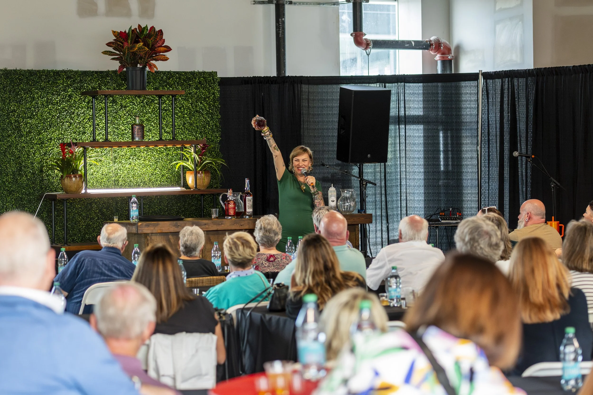 A woman in a green shirt is speaking at an event, holding a drink in one hand and a microphone in the other, with a lively audience seated in front of her.