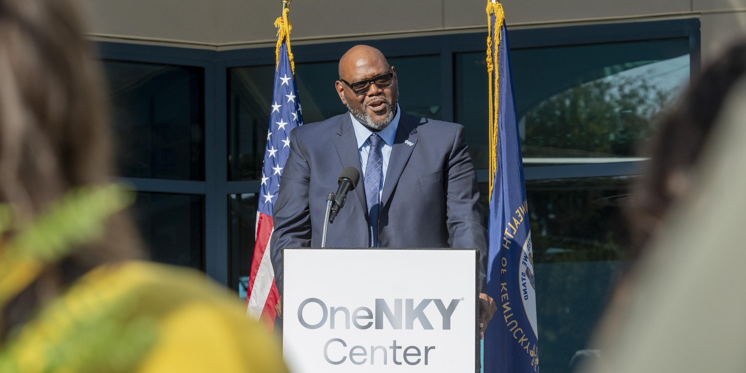 A man in a suit and sunglasses speaking at a podium with a microphone in front of an audience; there are American and Kentucky state flags behind him. The podium has a sign that reads 'OneNky Center'.