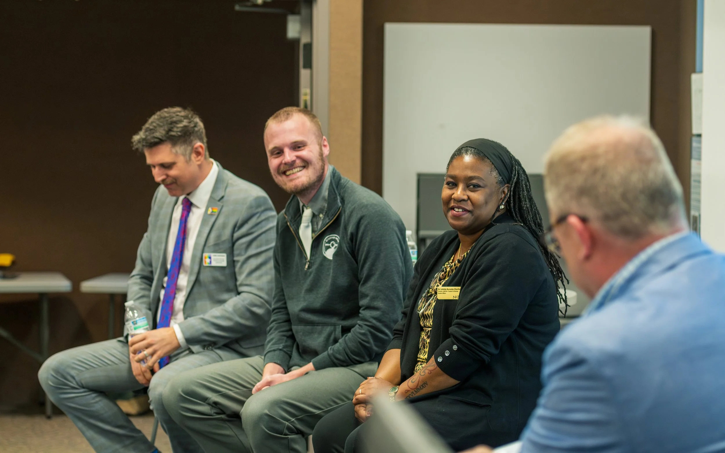 Four people sitting in a line at a panel discussion or meeting, including a man in a gray suit holding a water bottle, a smiling man with a beard wearing a gray jacket, a woman with dreadlocks in black clothing, and an older man with glasses in a blu
