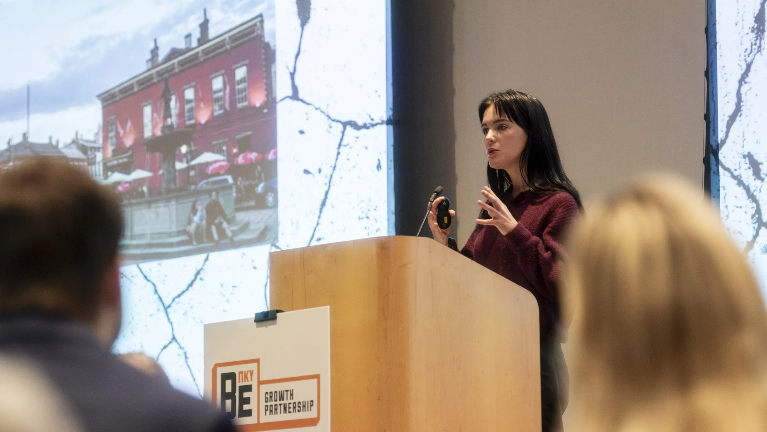 A woman giving a presentation at a conference, standing behind a wooden podium with a microphone, in front of a large screen displaying a photo of a red building with outdoor seating and umbrellas.