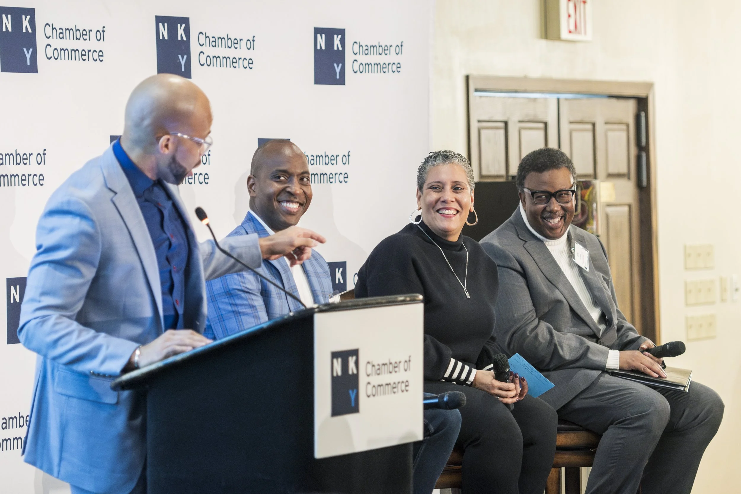 Four diverse professionals sitting on a panel at a chamber of commerce event, with one man standing at a podium and speaking.