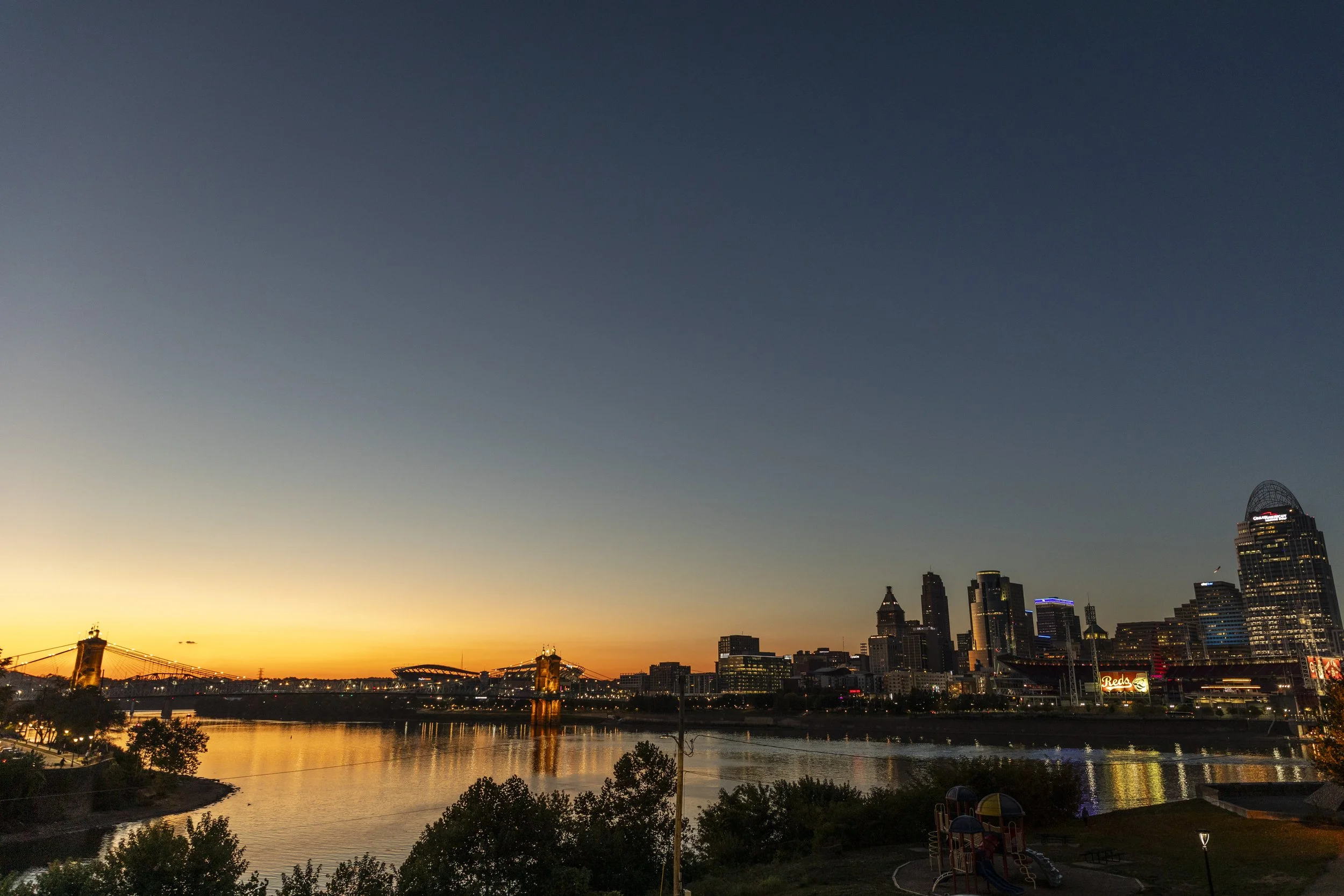 View of Cincinnati skyline at sunset with river in foreground and Columbus Bridge illuminated.