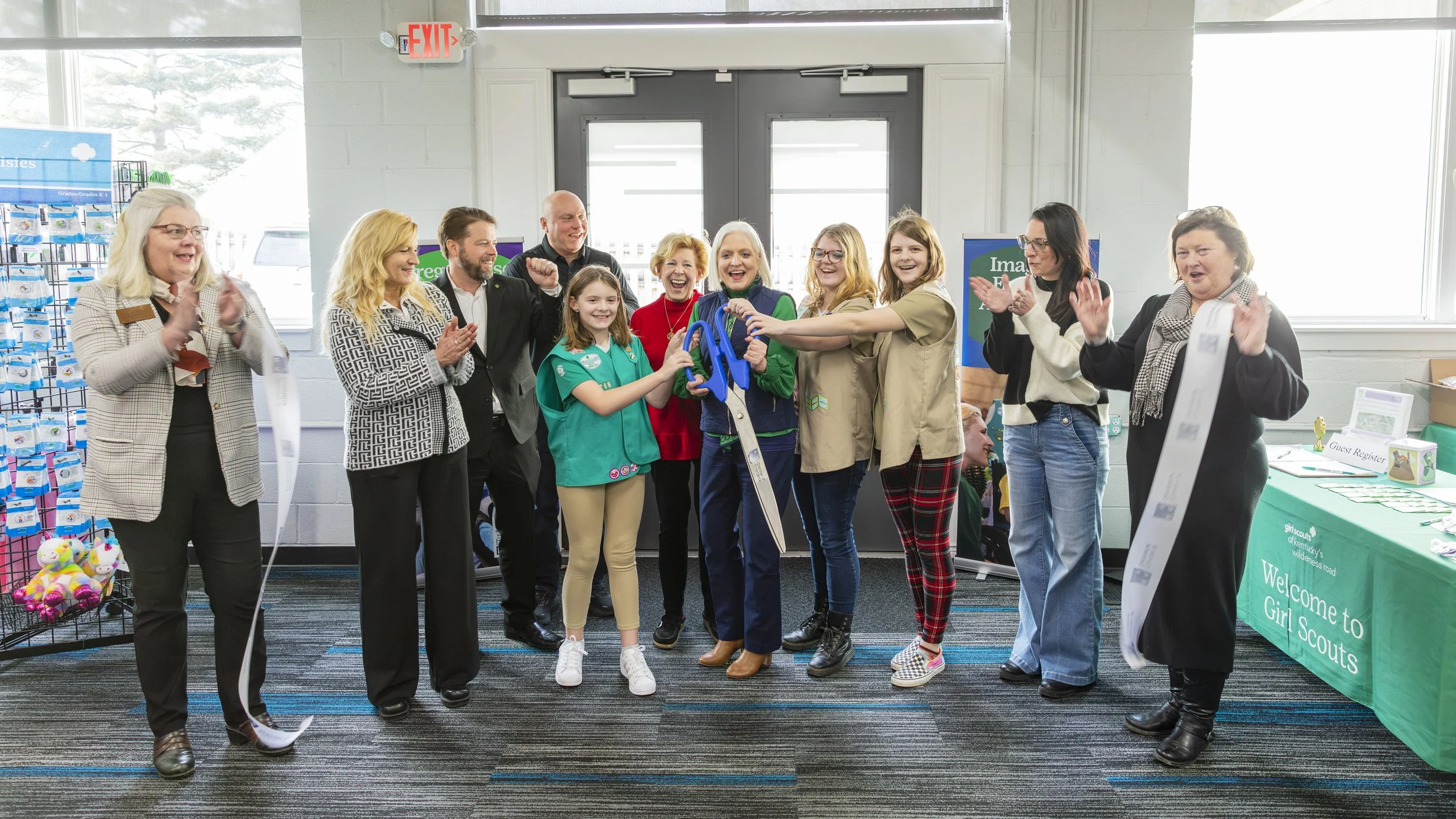 A group of people at a Girl Scouts event, with a woman holding large scissors about to cut a ribbon, surrounded by smiling attendees clapping and celebrating inside a room.