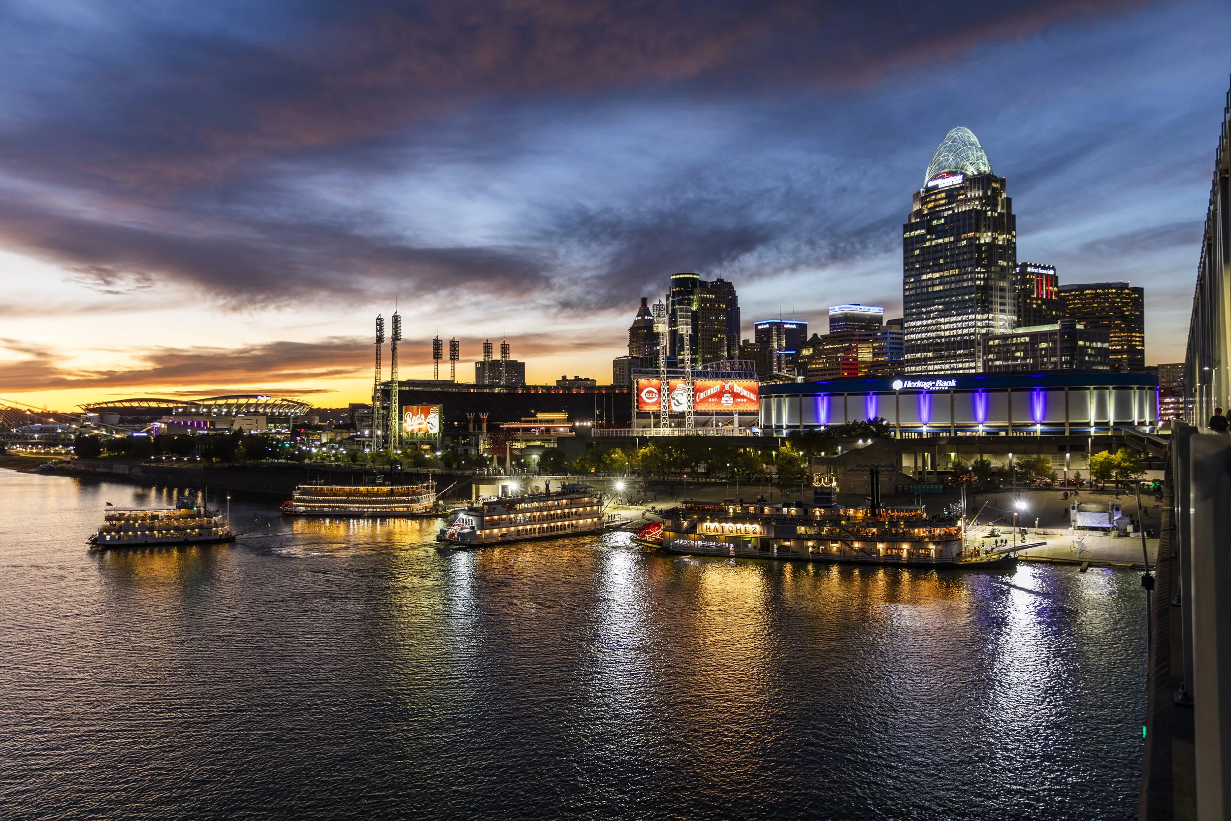 City skyline of Cincinnati at sunset, with colorful clouds, illuminated buildings, a river with boats, and reflections of city lights on the water.