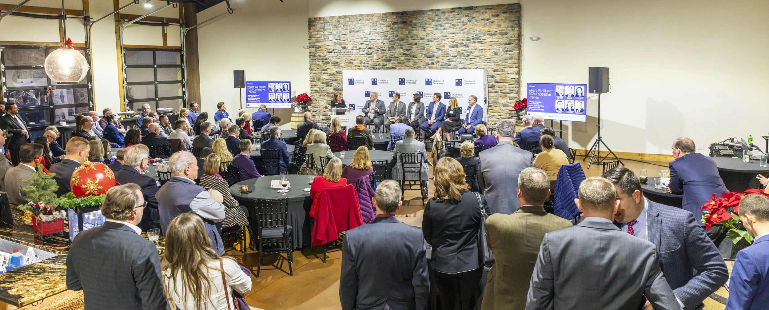 A large group of people attending a panel discussion inside a spacious room with a brick wall backdrop and large windows. The audience is seated at round tables, while a panel of speakers, including men and women in business attire, sit at a long tab