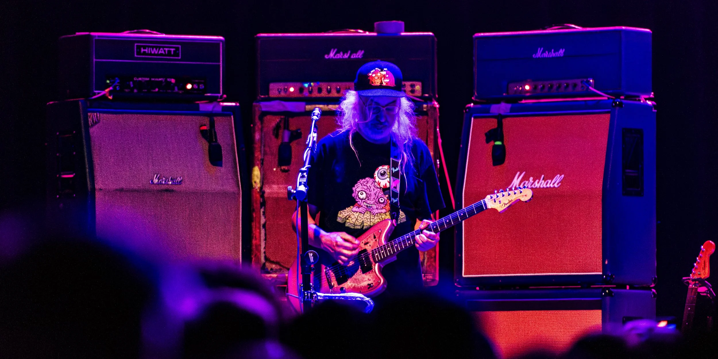 A musician with long hair wearing a black graphic t-shirt and a baseball cap, playing an electric guitar on stage with a dark background. Behind him are four large guitar amplifiers, including Marshall and Hiwatt.