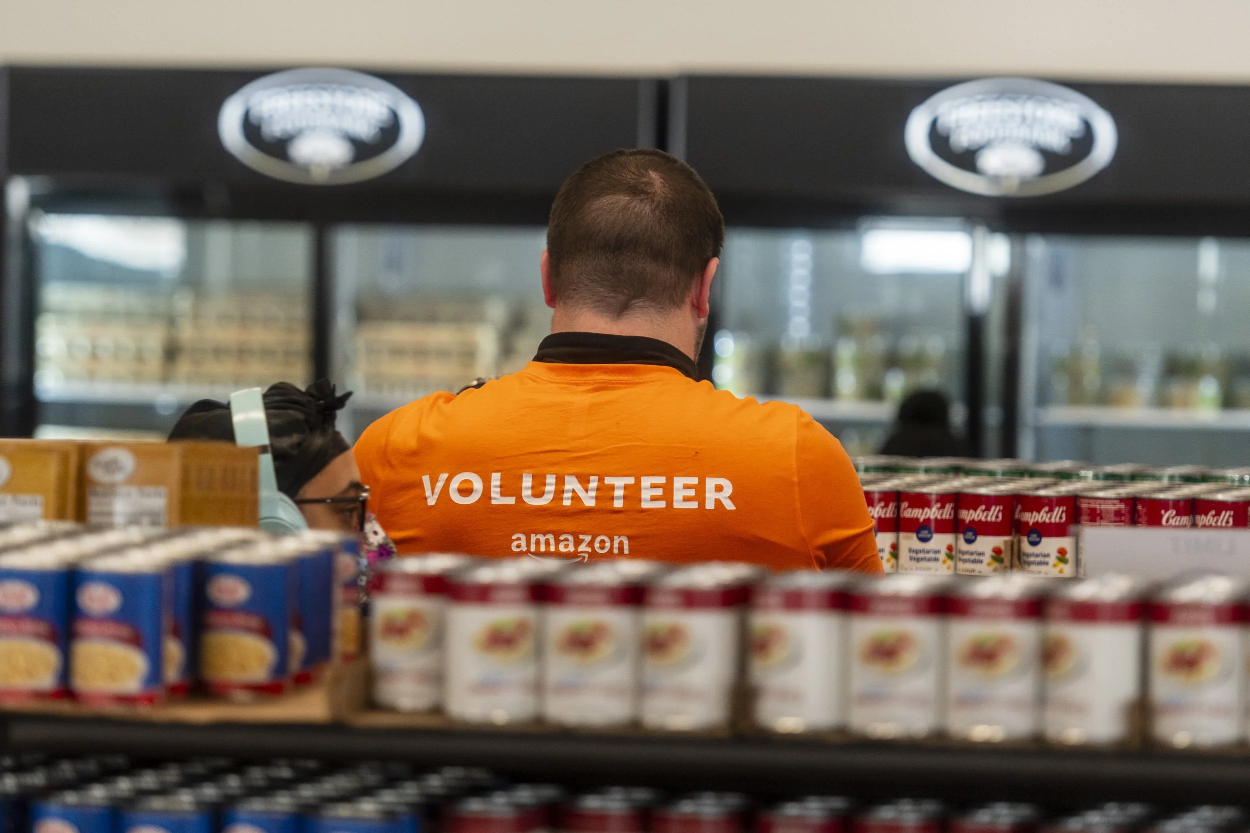 A volunteer wearing an orange t-shirt with 'VOLUNTEER' and 'amazon' printed on it, working in a grocery store aisle filled with canned goods.