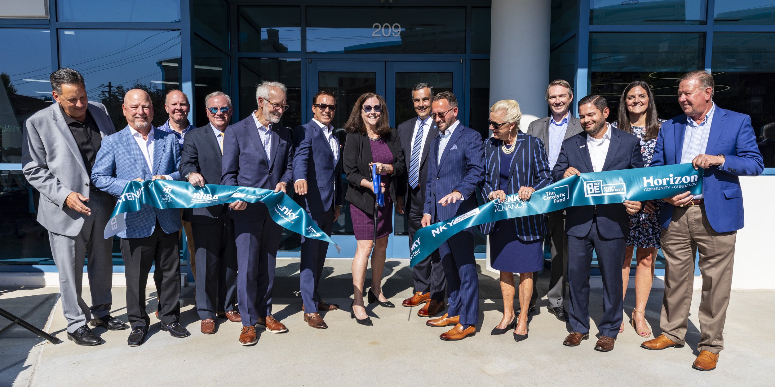 Group of people participating in a ribbon-cutting ceremony in front of a modern building with glass windows, with some individuals holding a blue ribbon and scissors.