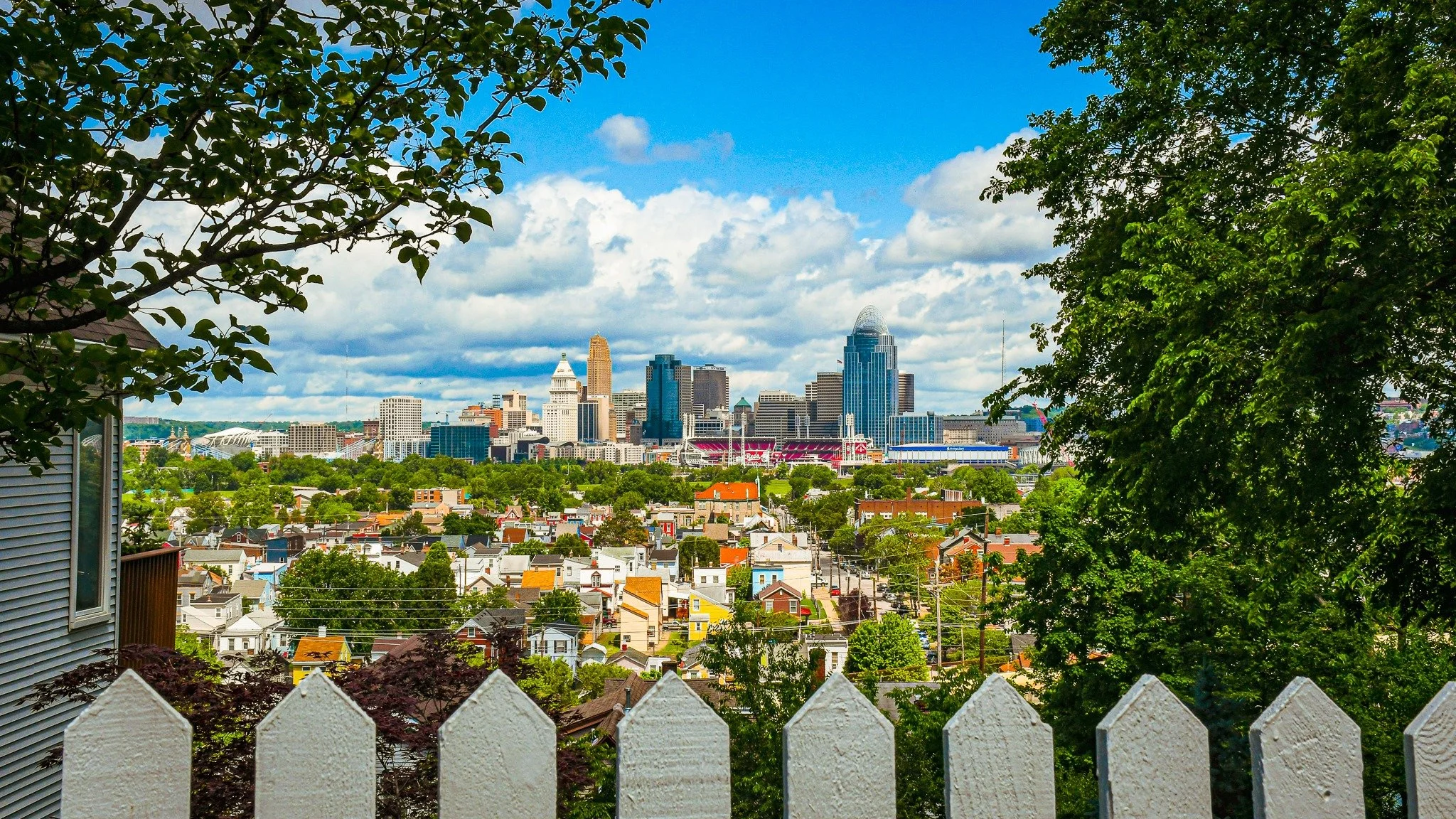 View of the Cincinnati skyline from a backyard, with trees and a white picket fence in the foreground on a sunny day with scattered clouds.