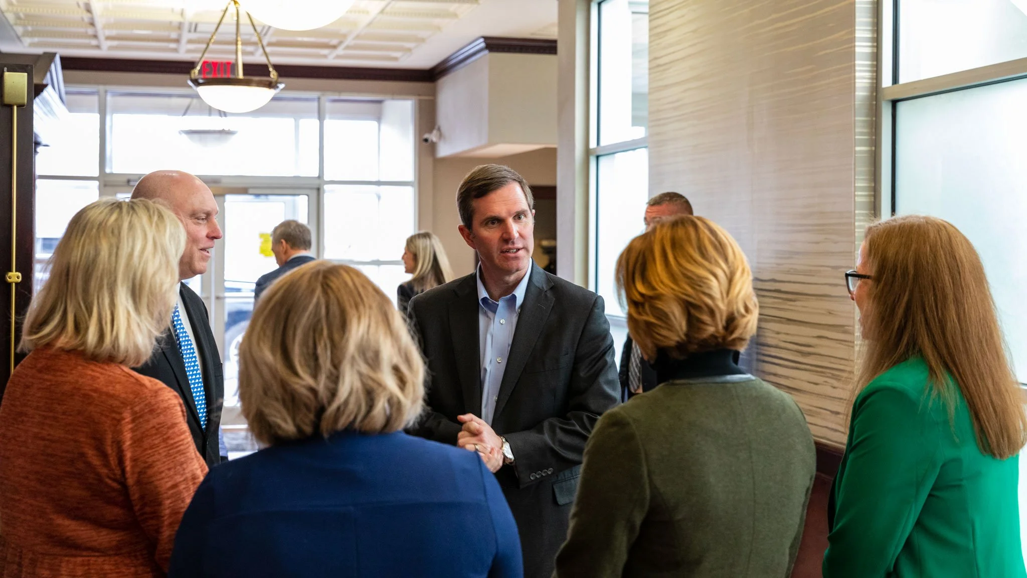 A man in a black suit speaking to a group of women and men in business attire in a room with large windows and a wooden wall.