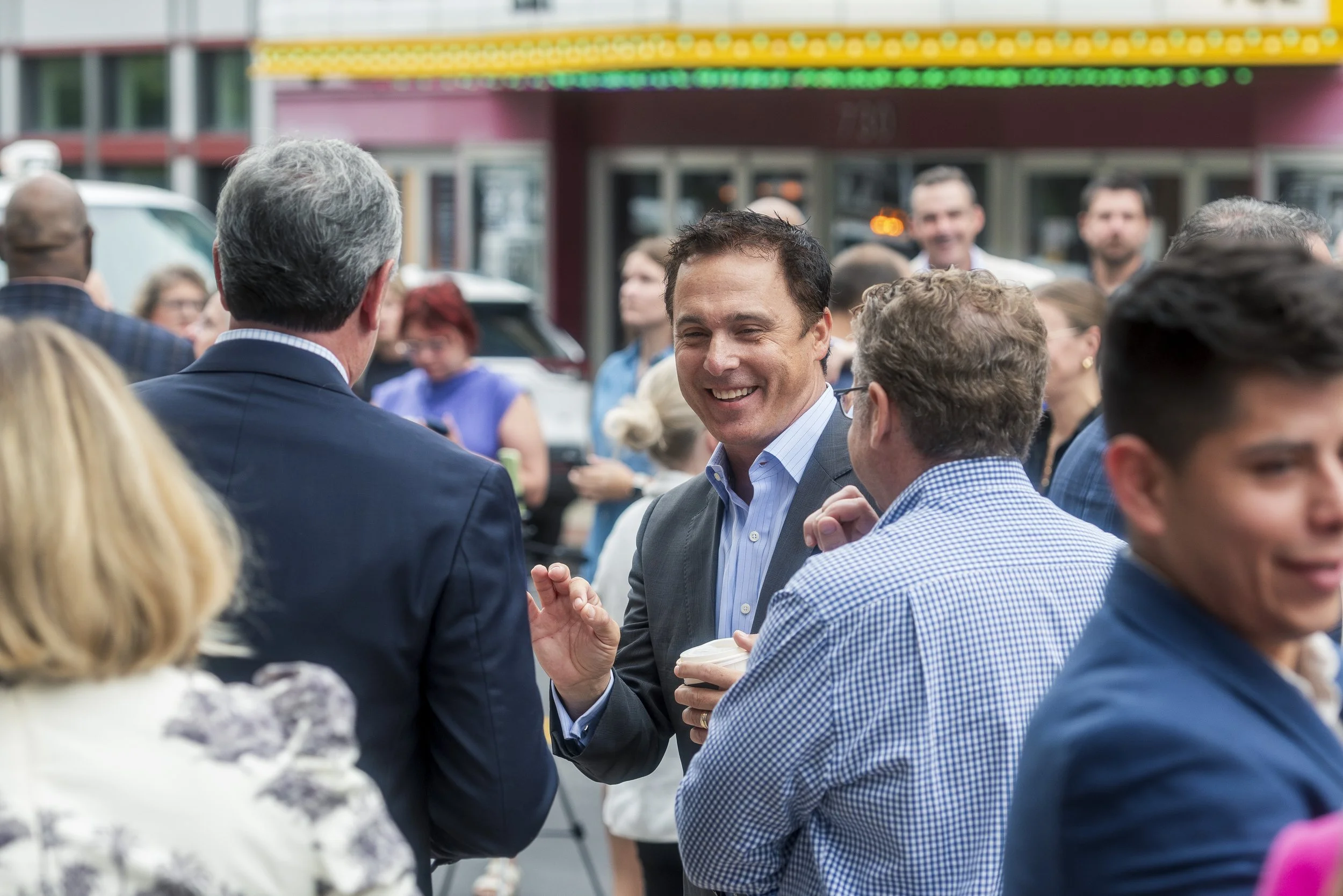 A group of people at an outdoor event, engaging in conversation. In focus is a man smiling and holding a coffee cup, wearing a light blue shirt and gray blazer, surrounded by others in business and casual attire.