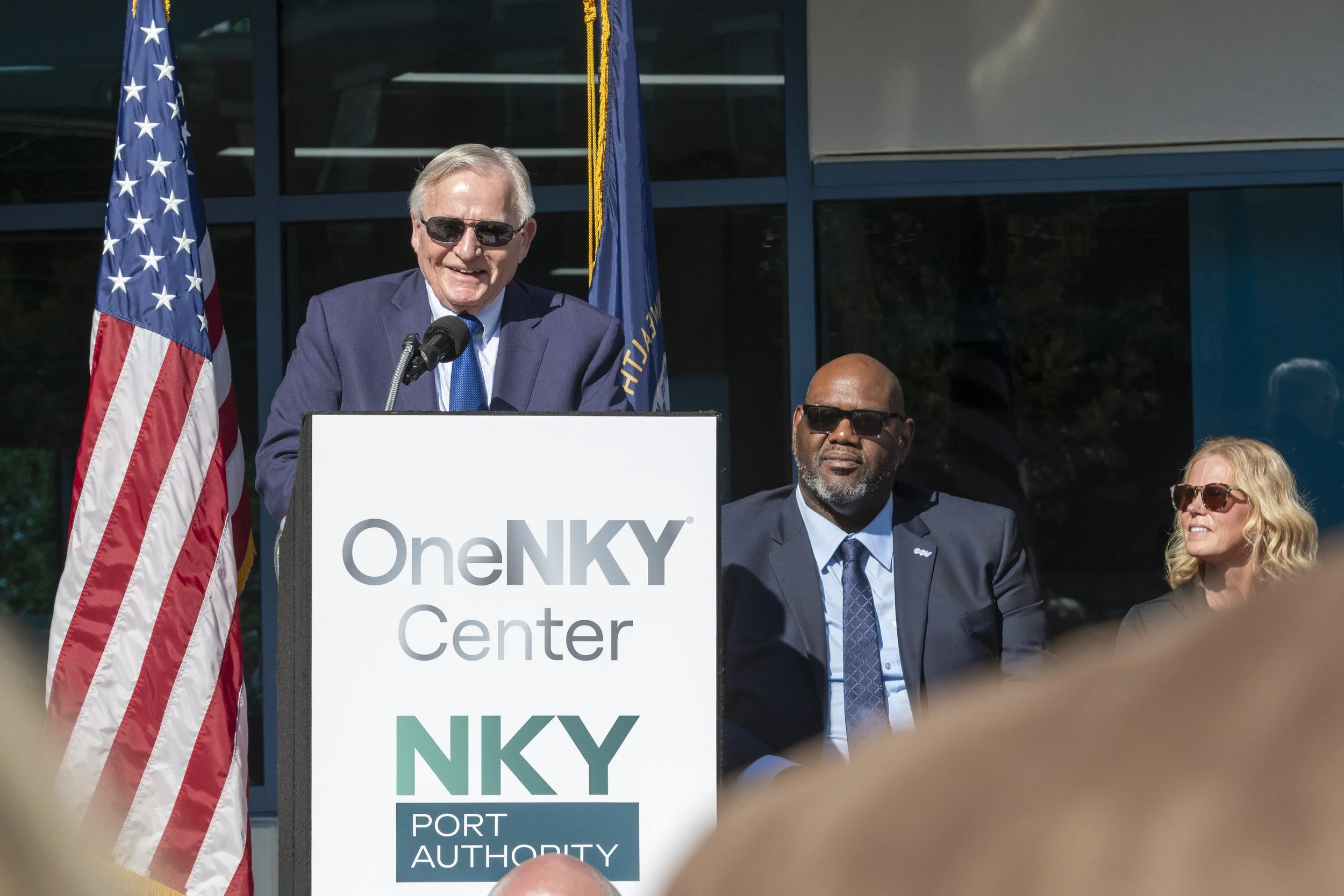 A man in a blue suit and sunglasses is speaking at a podium with a sign that reads "OneNKY Center" and "NKY Port Authority". Two people, a man and a woman, are seated nearby, also wearing suits and sunglasses. An American flag and a blue flag are beh