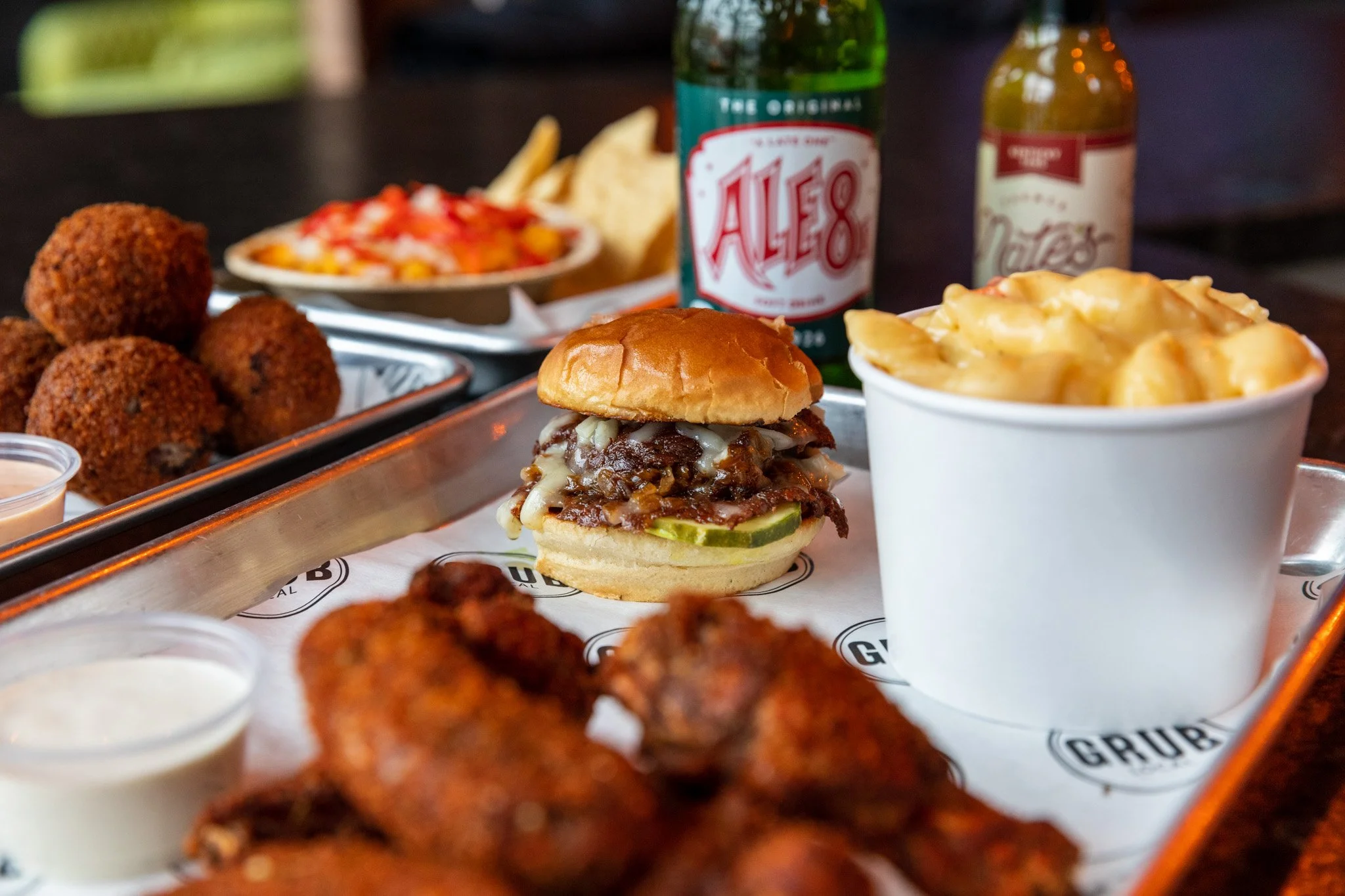 A tray featuring a cheeseburger with pickles and melted cheese, a serving of macaroni and cheese, fried chicken wings, croquettes, a small container of dressing, a bottle of Ale 8 soda, and a bottle of honey mustard sauce on a wooden table.