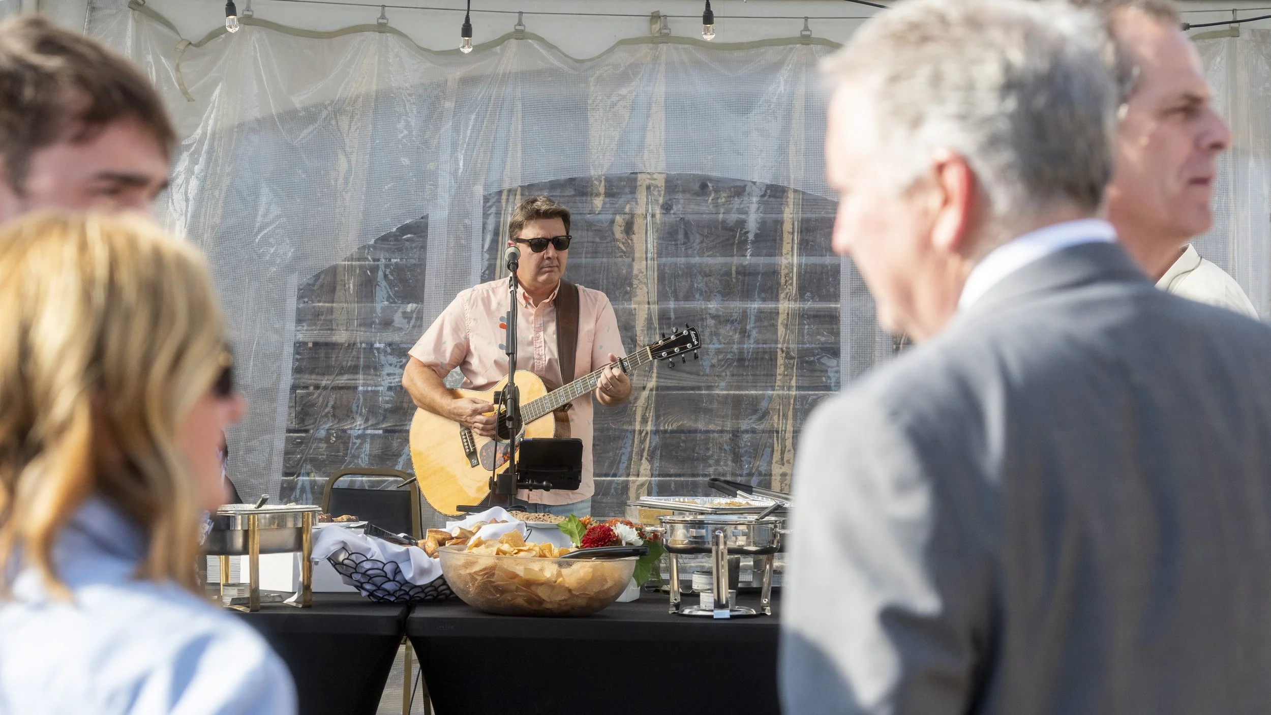 A man wearing sunglasses and a pink shirt is playing an acoustic guitar and singing into a microphone at a daytime outdoor event, with a buffet table in front of him and people in the foreground.