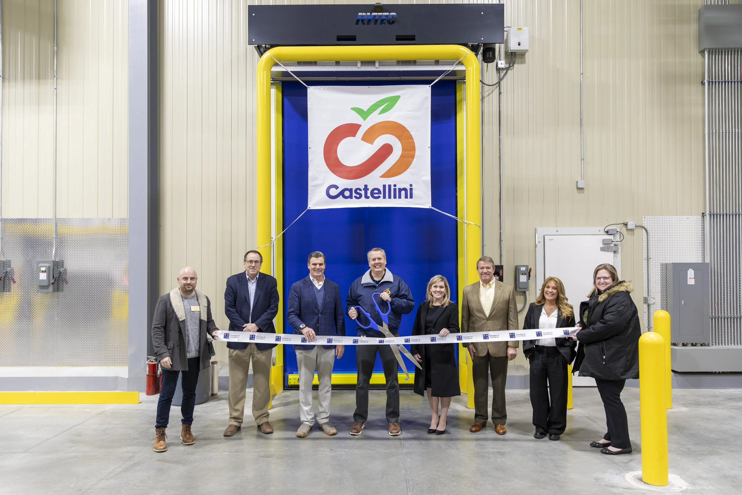 Group of people standing in front of a large folding door with Castellini logo, participating in a ribbon-cutting event, inside an industrial warehouse with beige and metallic walls.