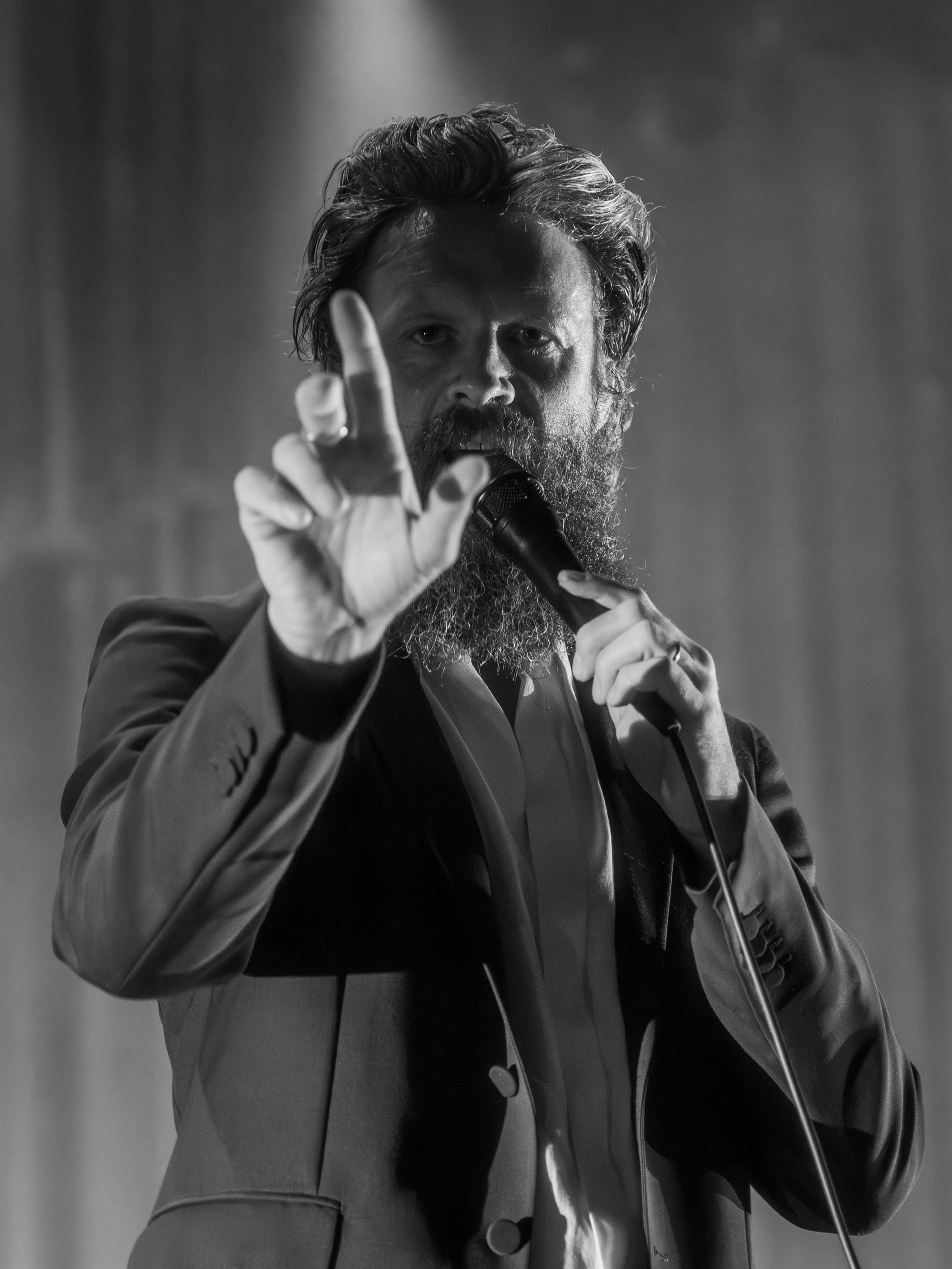 A man with a beard and styled hair, dressed in a suit, speaking into a microphone while gesturing with his hand during a performance or speech, in black and white.