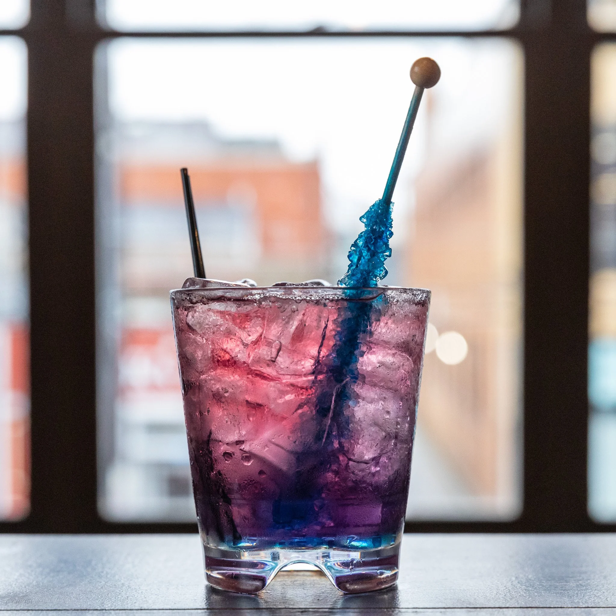 A glass filled with a pinkish-purple cocktail, ice, and a blue stir stick with crushed blue candy on it, set against a window background.