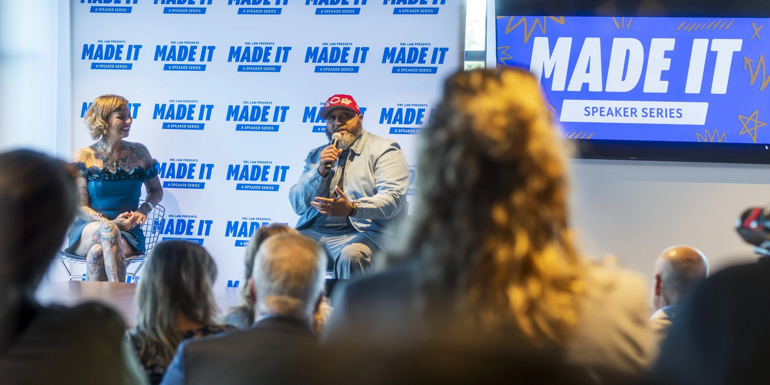 A panel discussion at an event titled 'Made It', with one woman and one man on stage. The woman has tattoos and is smiling, and the man is speaking into a microphone. The background features a large banner and a screen with the event logo.