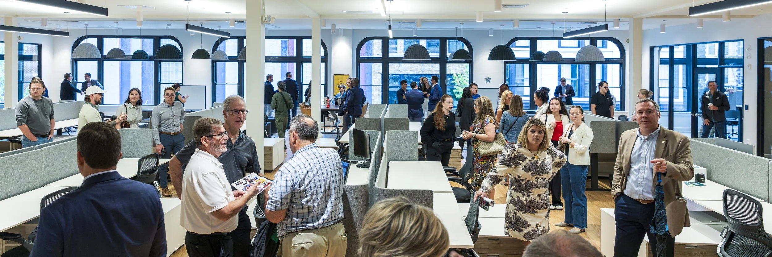 A busy modern office space filled with people engaging in conversations. The room has large windows, black pendant lights, and a mix of open seating and standing areas.