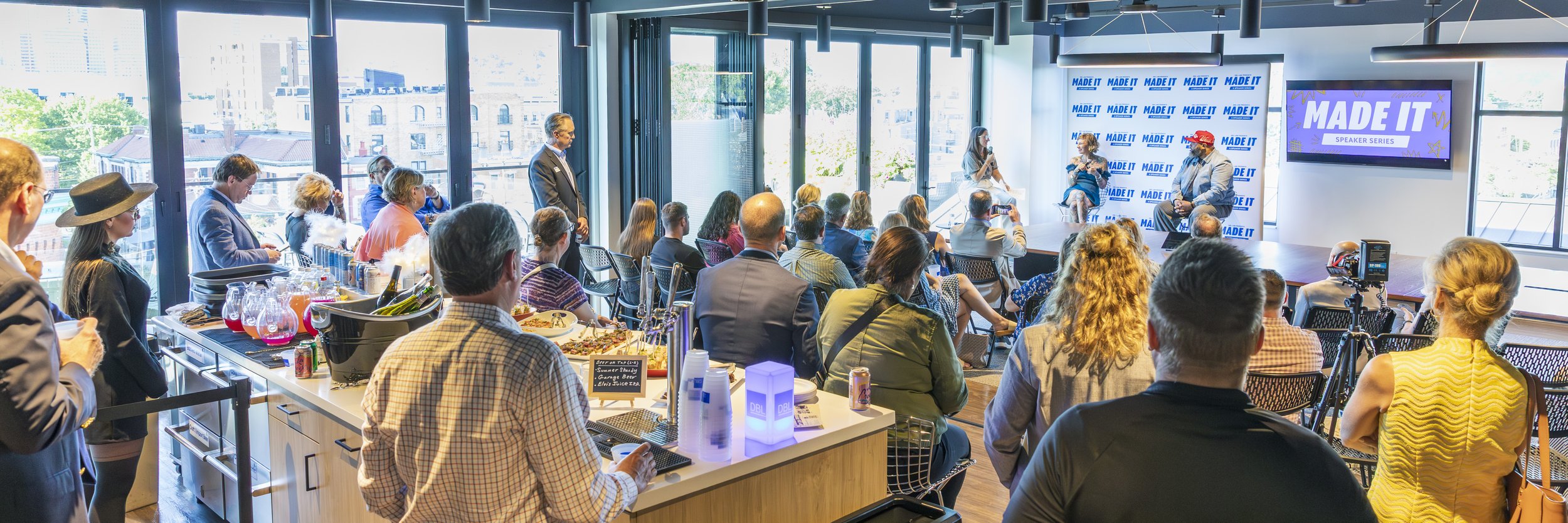 A conference or talk event taking place in a modern, bright room with large windows. Audience members sitting and standing, listening to two women on stage, one speaking and one seated. A large screen displays 'MADE IT', and a branding backdrop with 