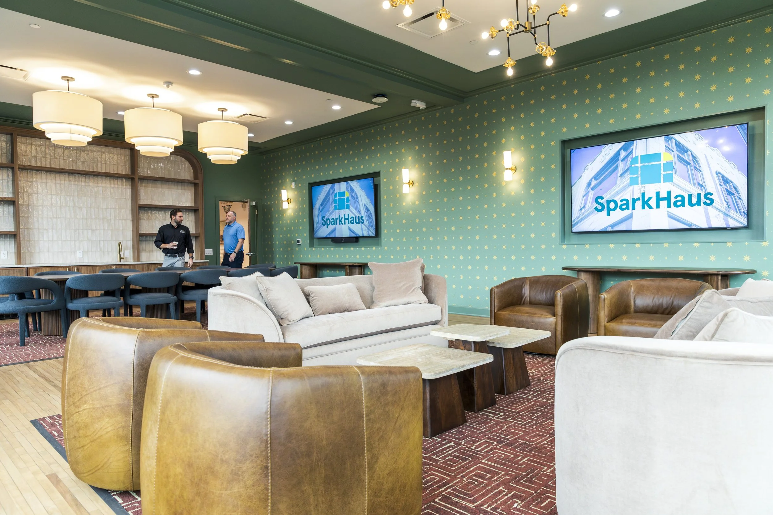 A spacious lounge area with green walls decorated with gold star patterns, multiple television screens displaying 'SparkHaus' branding, and comfortable seating including sofas and leather armchairs. Two men are standing and talking near a bar area in
