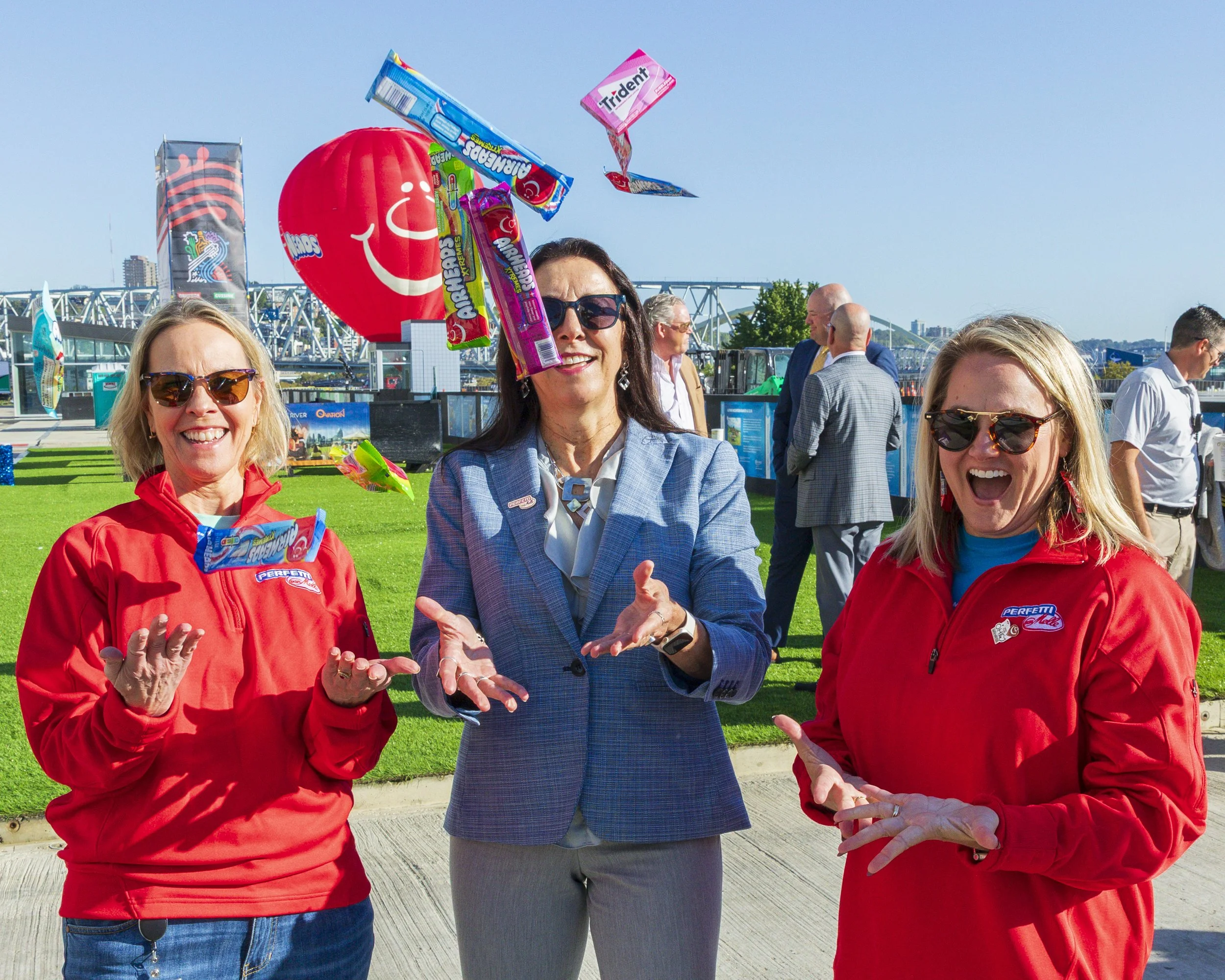Three smiling women in sunglasses outdoors on a sunny day, two wearing red jackets and one in a light blue blazer, in a park or event area with a grassy background, colorful candy being thrown in the air, and a large red smiley face balloon in the ba