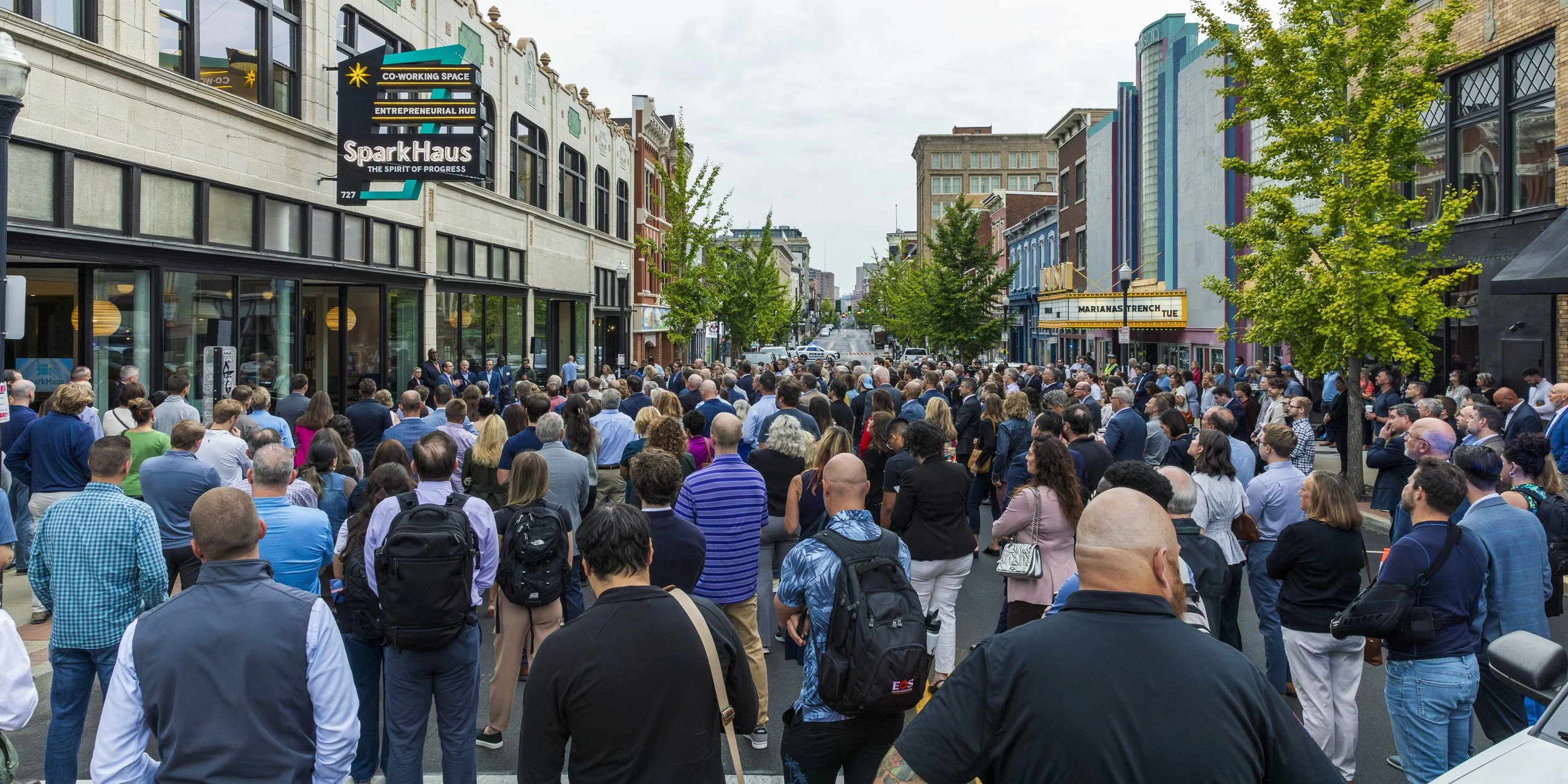 Crowd of people gathered outdoors on a city street, attending an event or protest, with buildings and trees visible on both sides.