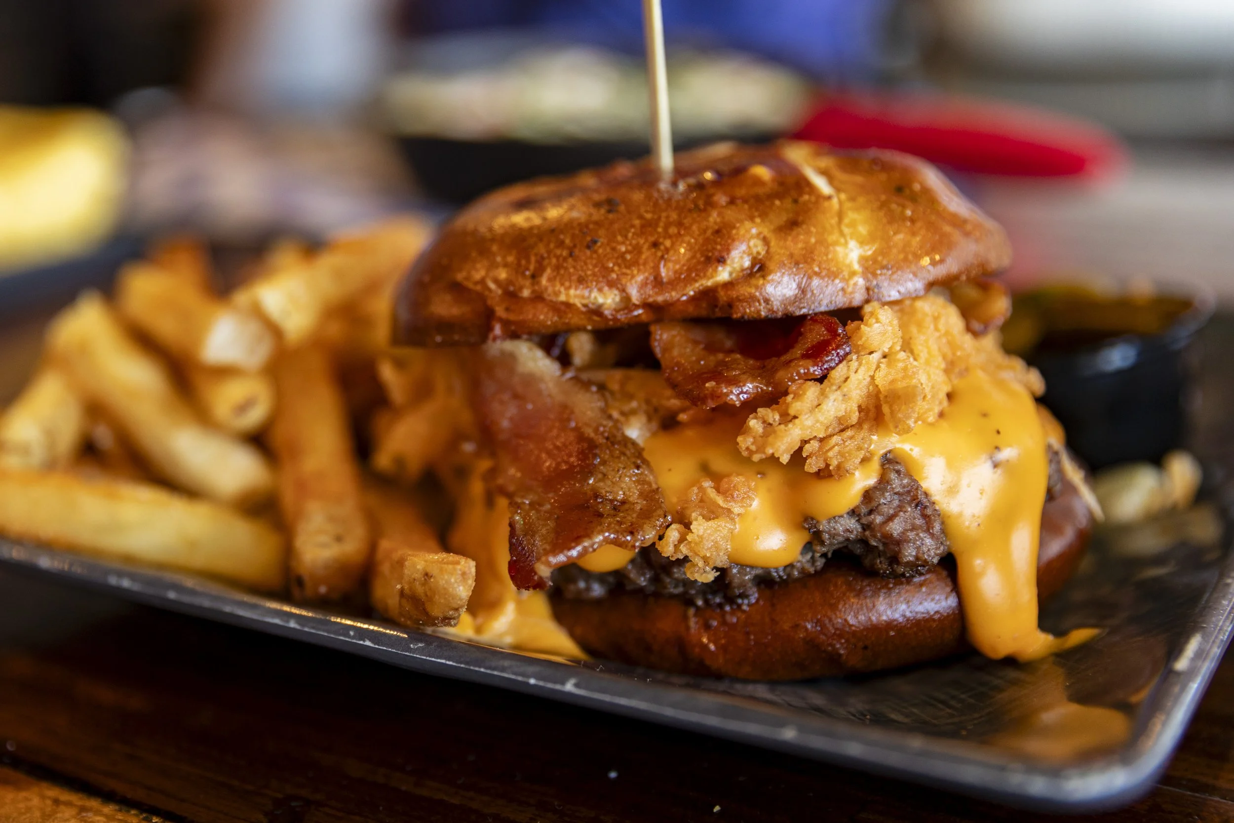 Close-up of a cheeseburger with bacon and fried onion rings, served with French fries on a black tray.