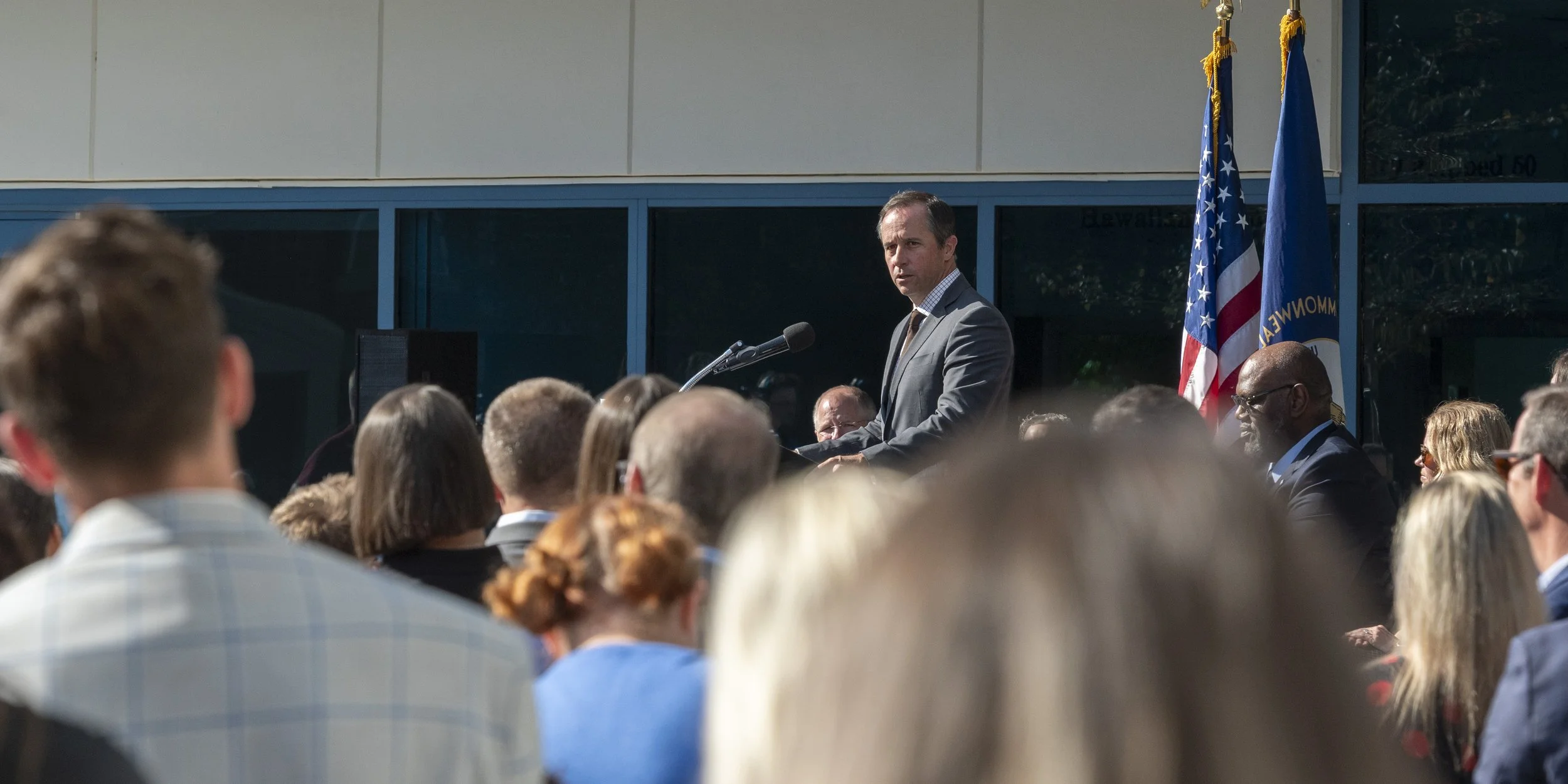 A man in a suit delivering a speech at an outdoor event with a crowd, with an American flag and another flag behind him.