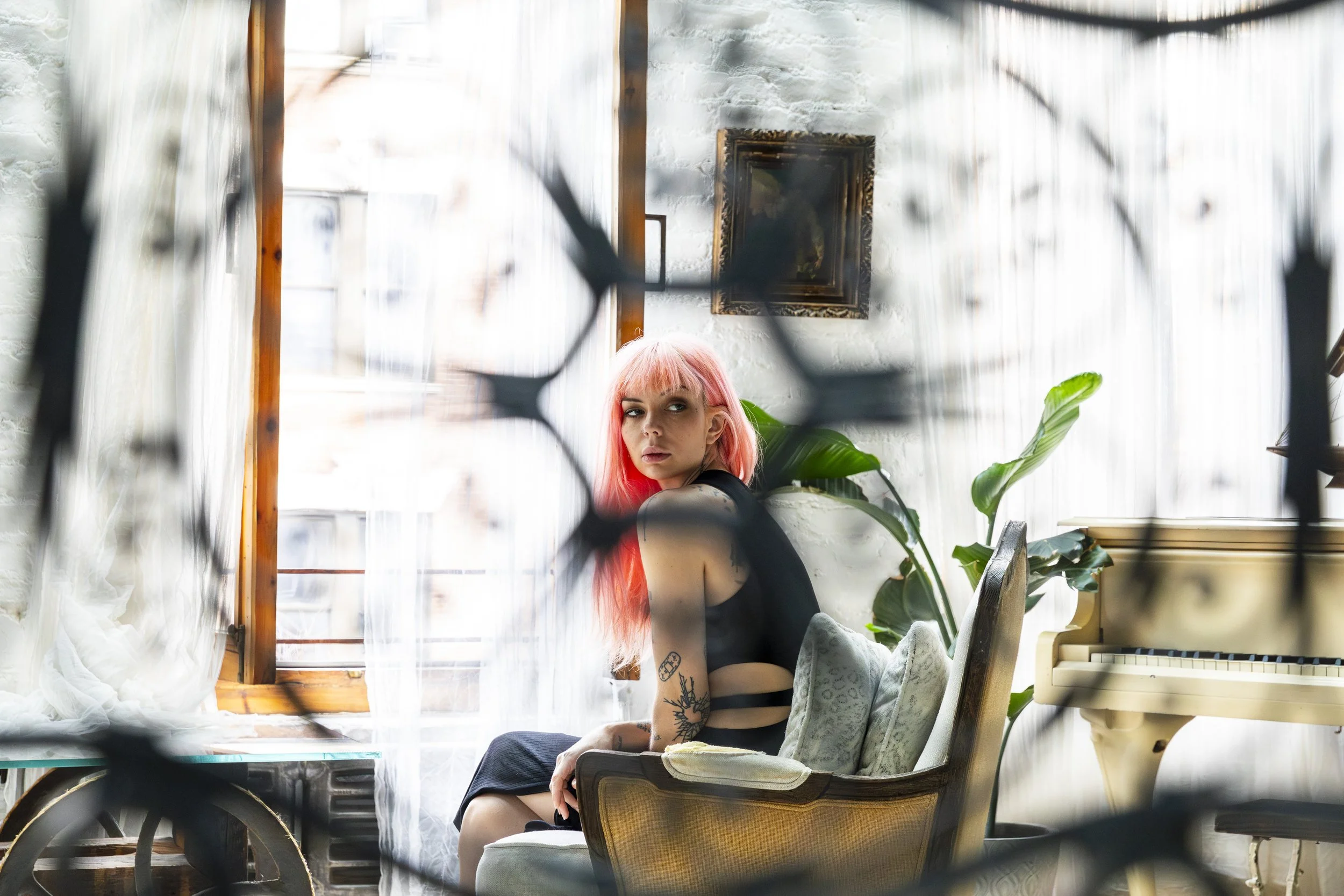 A woman with pink hair sitting on a vintage armchair inside a room, framed by dark branches in the foreground. She is looking towards the camera with a serious expression, wearing a black sleeveless top, and has tattoos on her arms. The room has whit